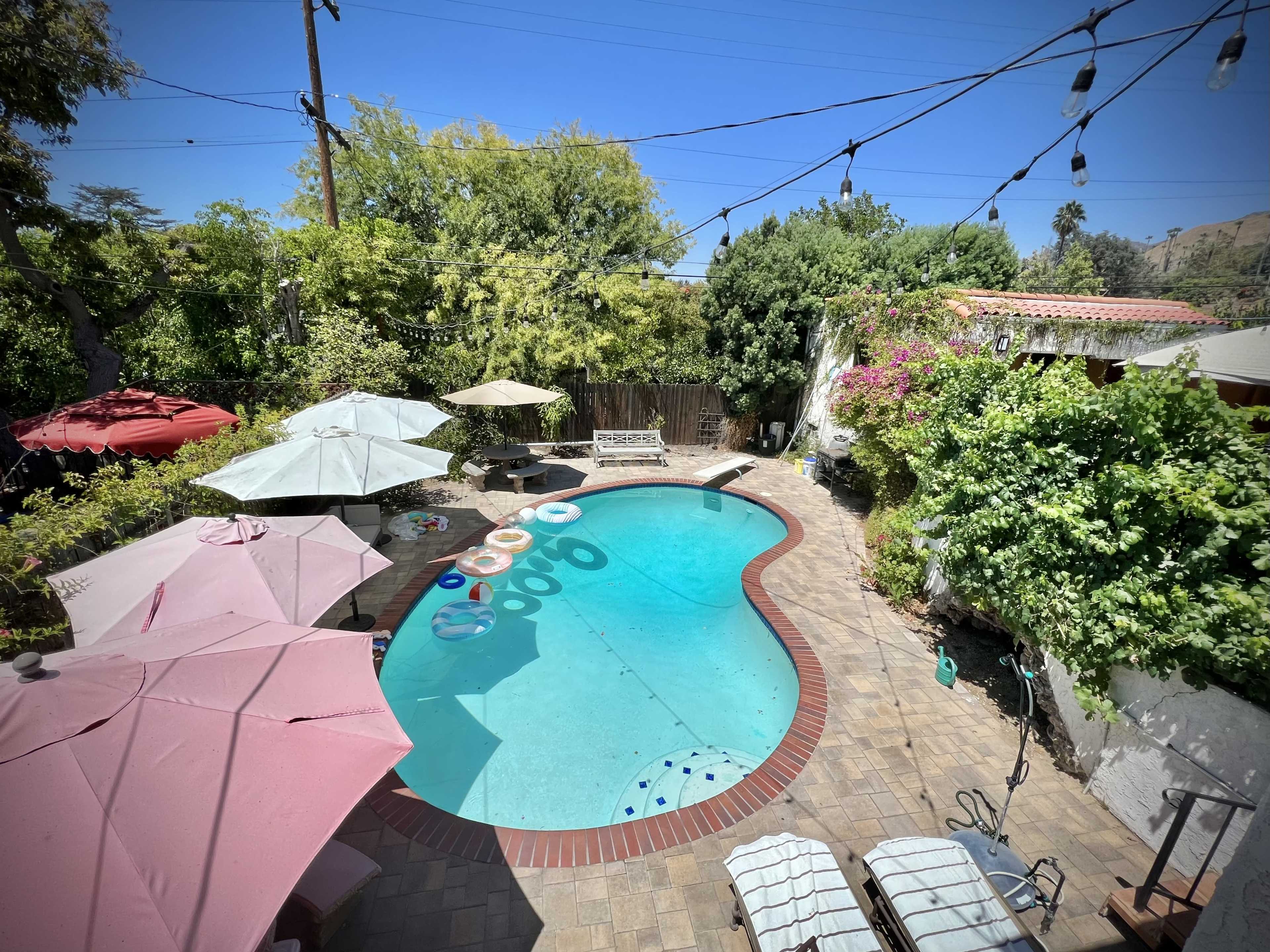 A backyard swimming pool surrounded by lush greenery, patio umbrellas, and floating pool toys.