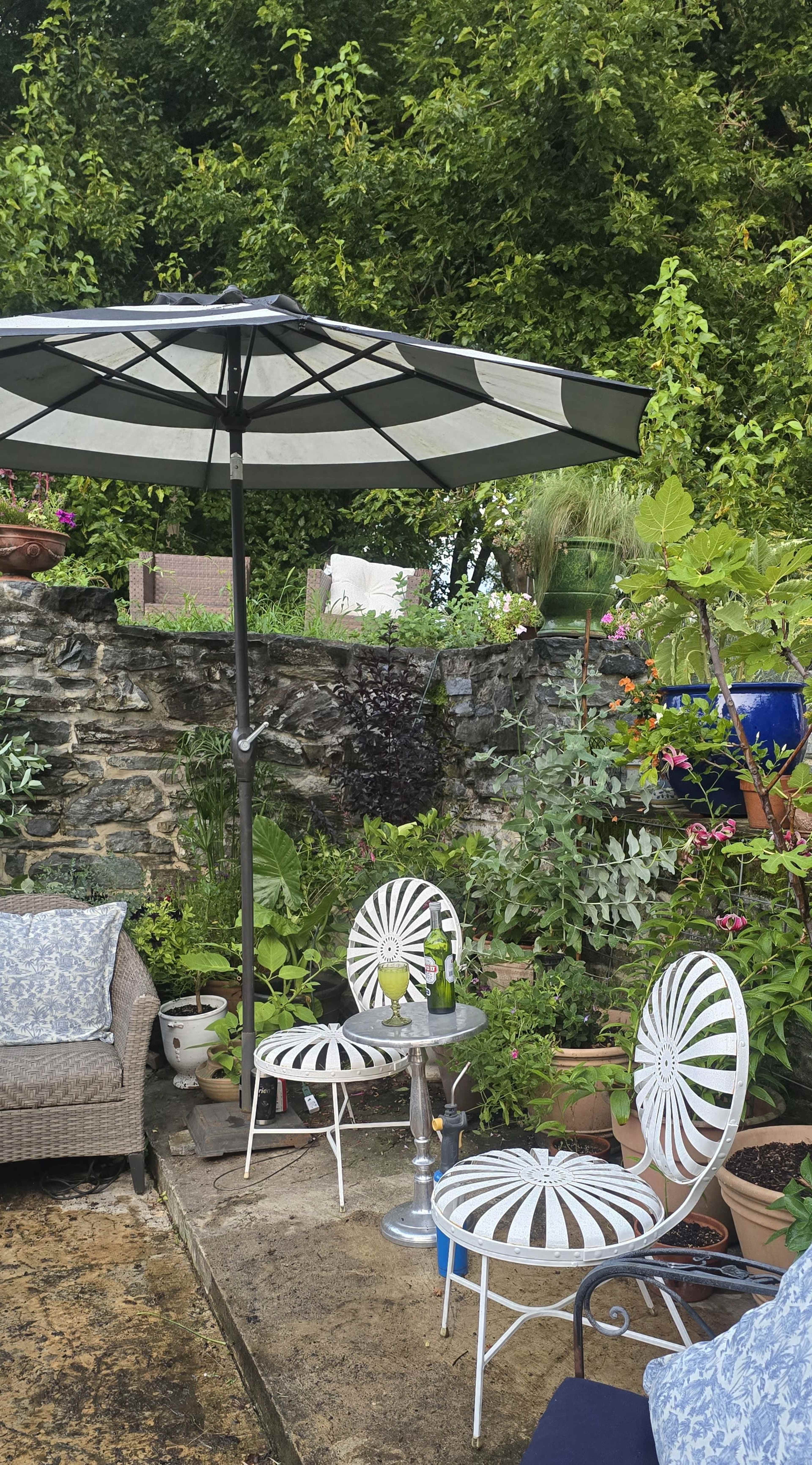 A small outdoor seating area features two striped chairs, a table, and an umbrella surrounded by various potted plants and a stone wall.