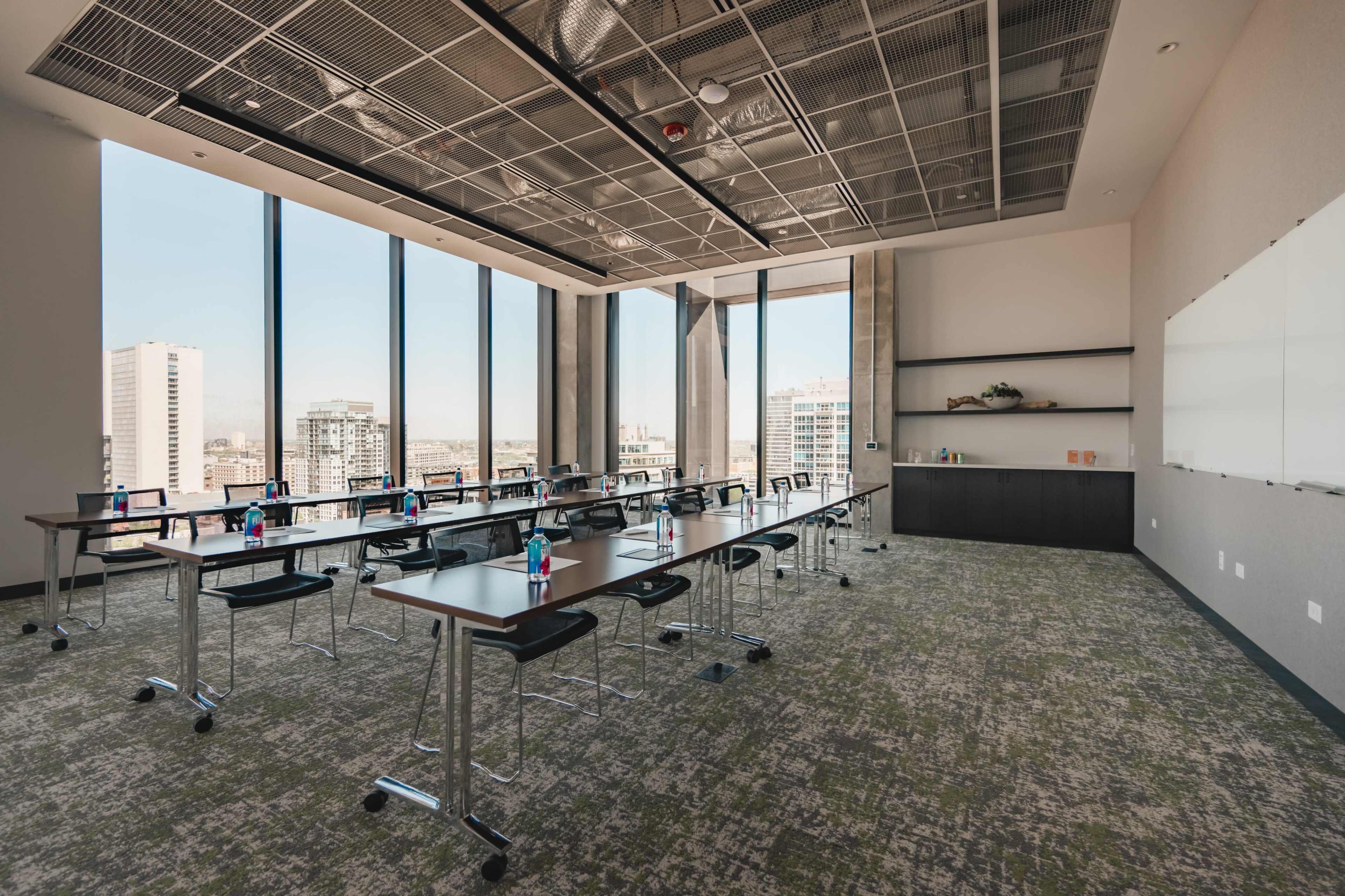 The image shows a modern conference room with large windows, arranged with several tables and chairs, and equipped with a whiteboard and a shelf in one corner.