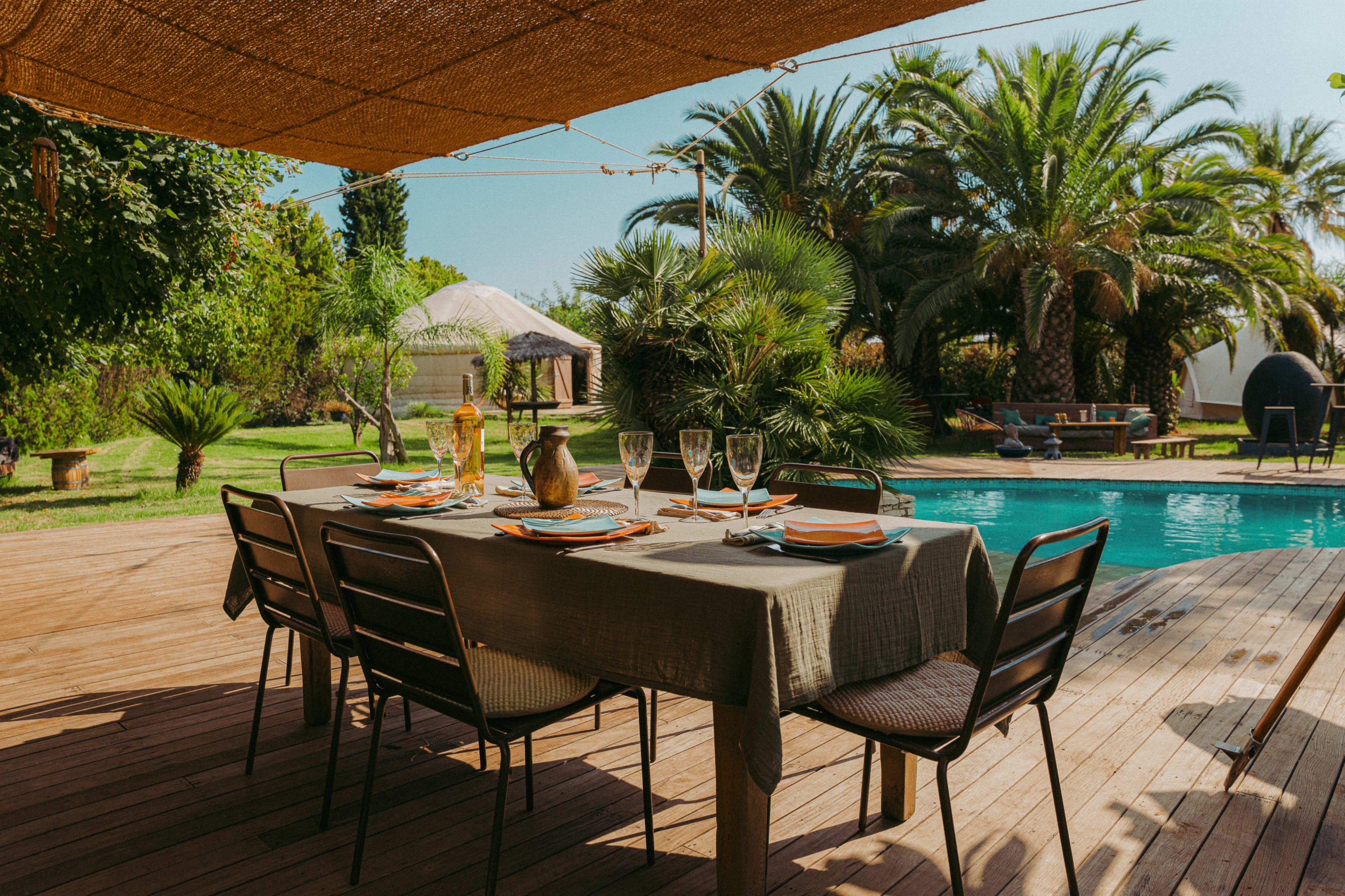 A outdoor dining table is set beside a pool, surrounded by lush palm trees and garden greenery.