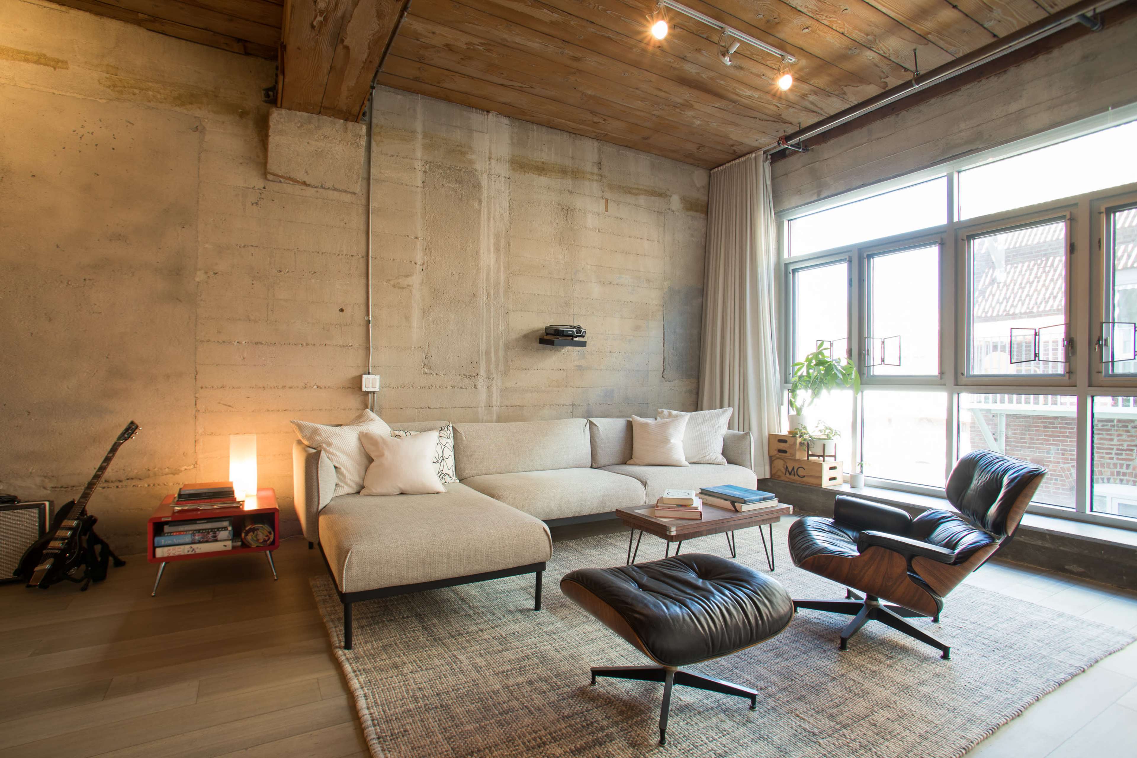 The image shows a modern, minimalistic living room with a sofa, an armchair, a coffee table, and large windows allowing natural light to enter.