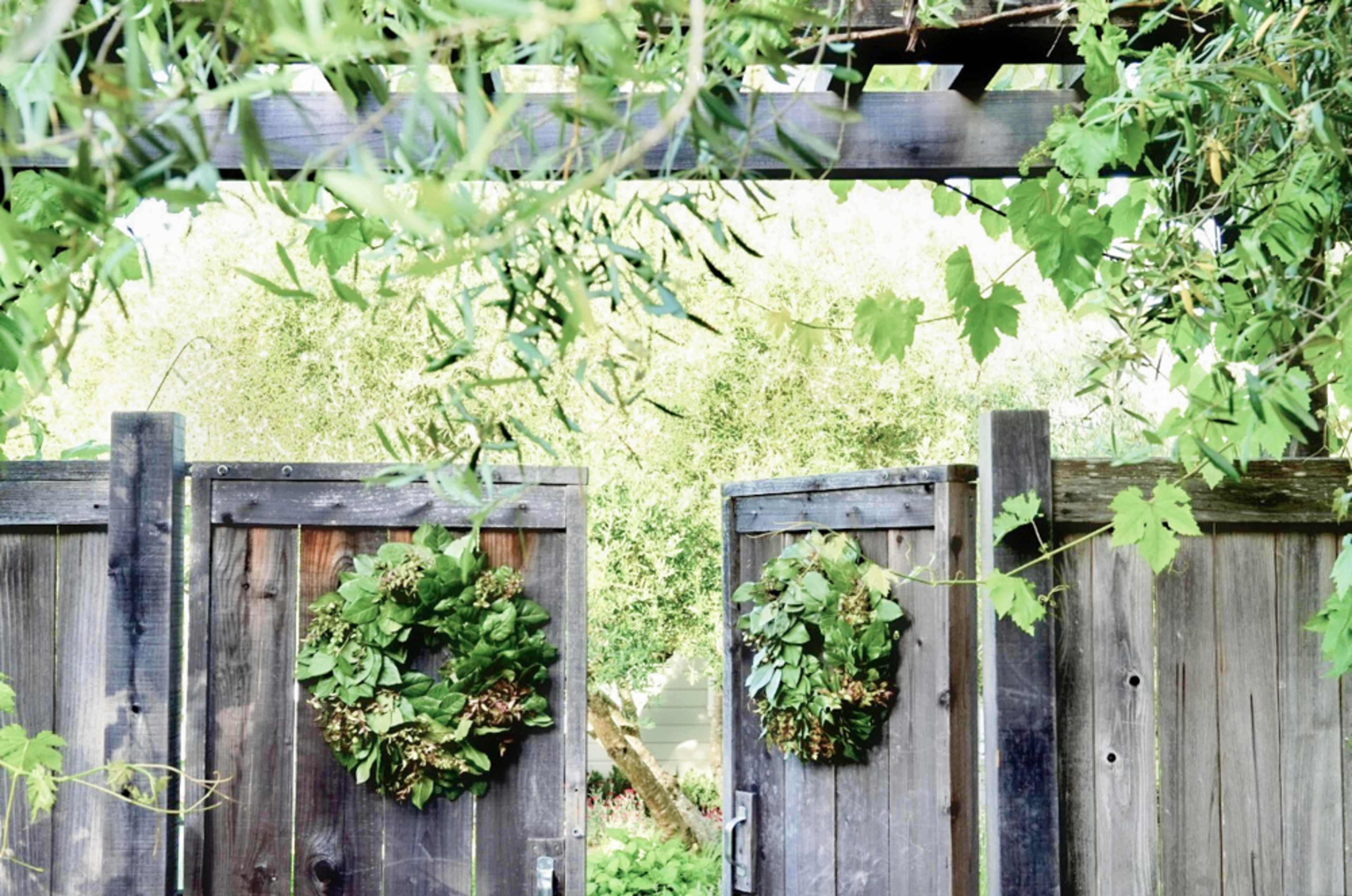 Two wooden gates adorned with wreaths of greenery are partially open, leading into a lush garden beyond.