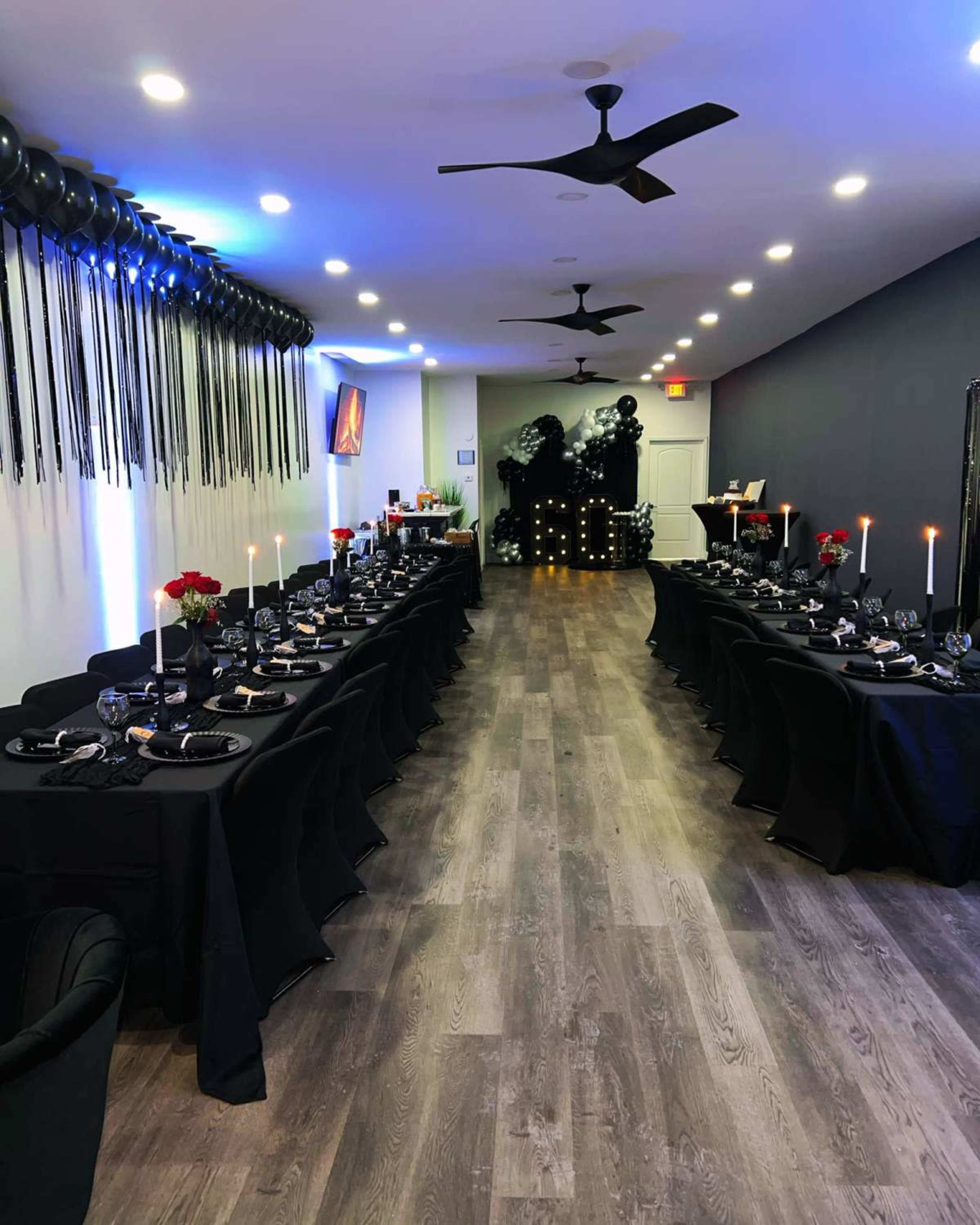 A long, elegantly set dining table with black tablecloths is arranged in a dimly lit room, featuring decorative candles, black balloons, and a wall adorned with dark accents.