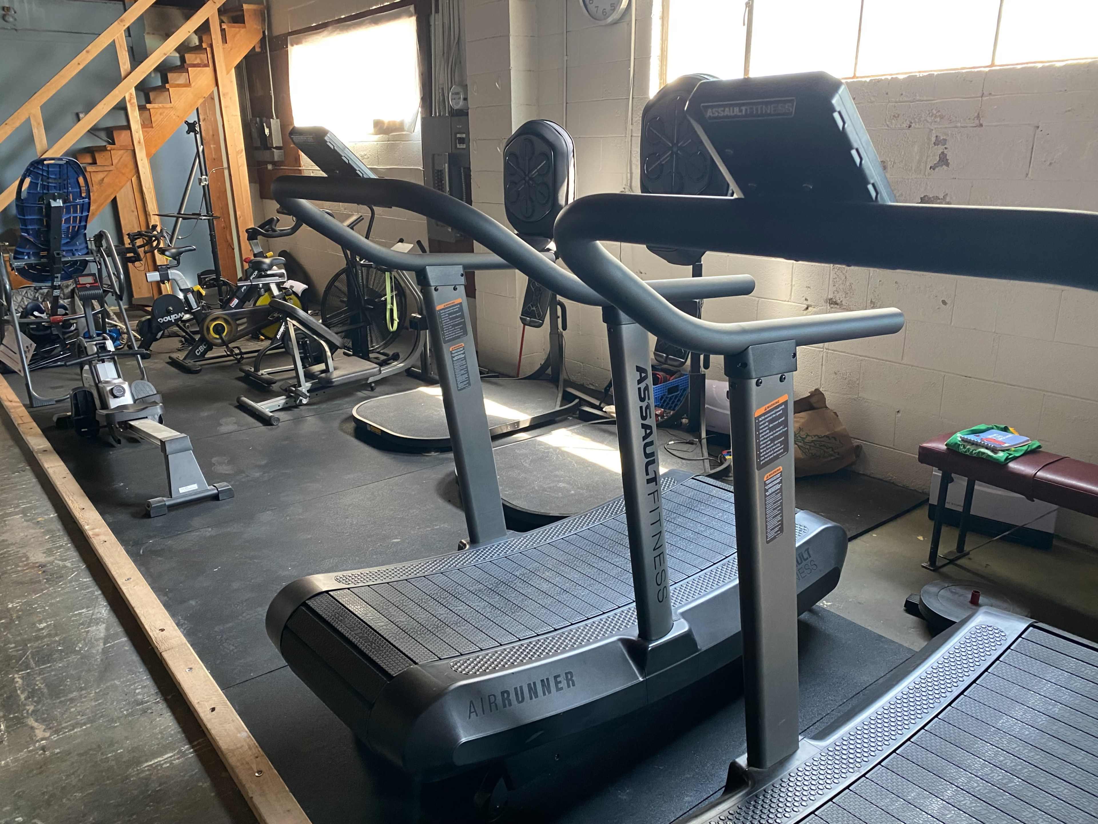 A gym space featuring several fitness machines, including two curved treadmills and various stationary bikes, along with a stairway in the background.