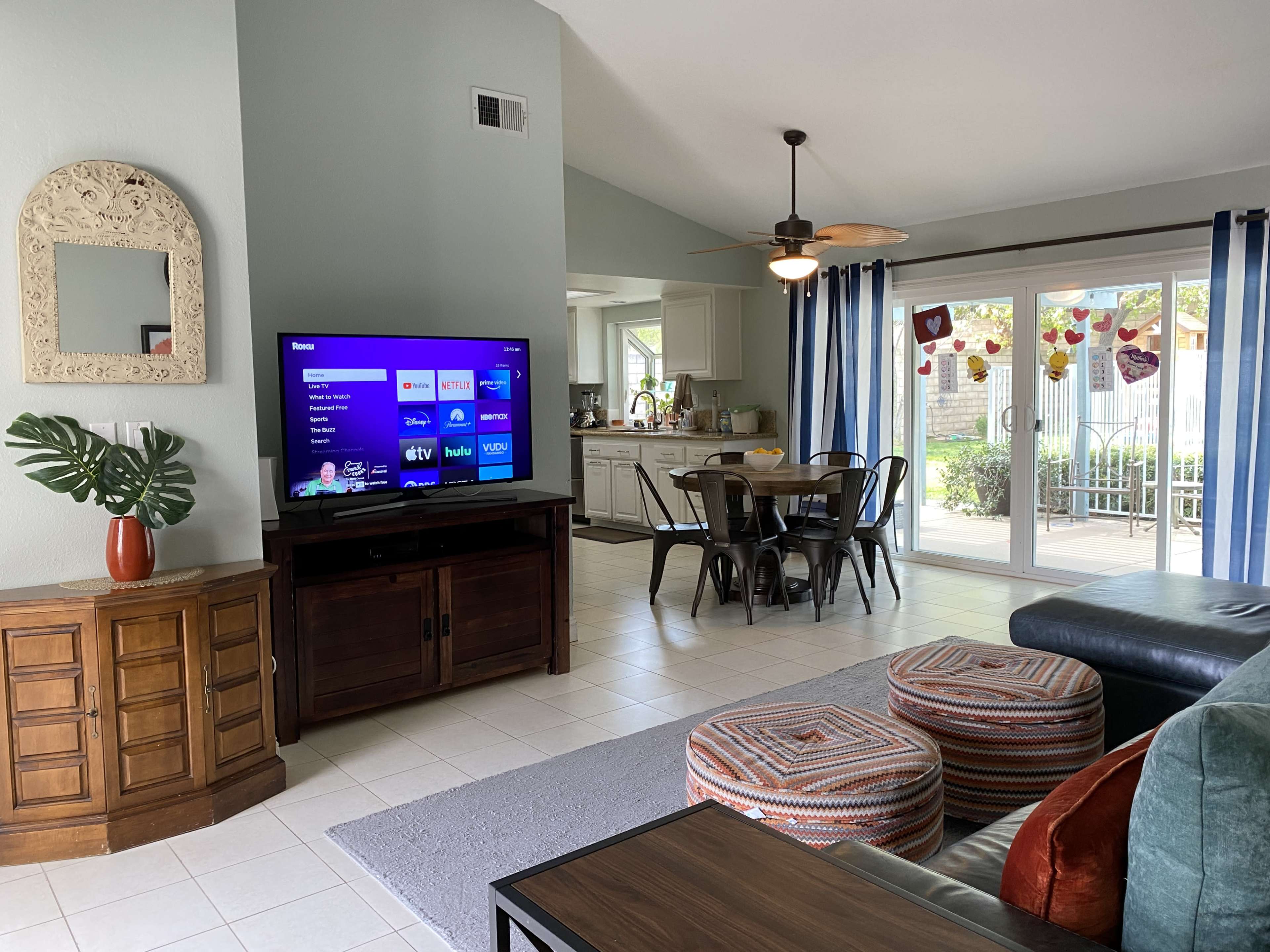 A living room with a large TV on a wooden cabinet, a sofa with patterned cushions, and a dining area with a round table and chairs near a glass door leading to an outdoor space.