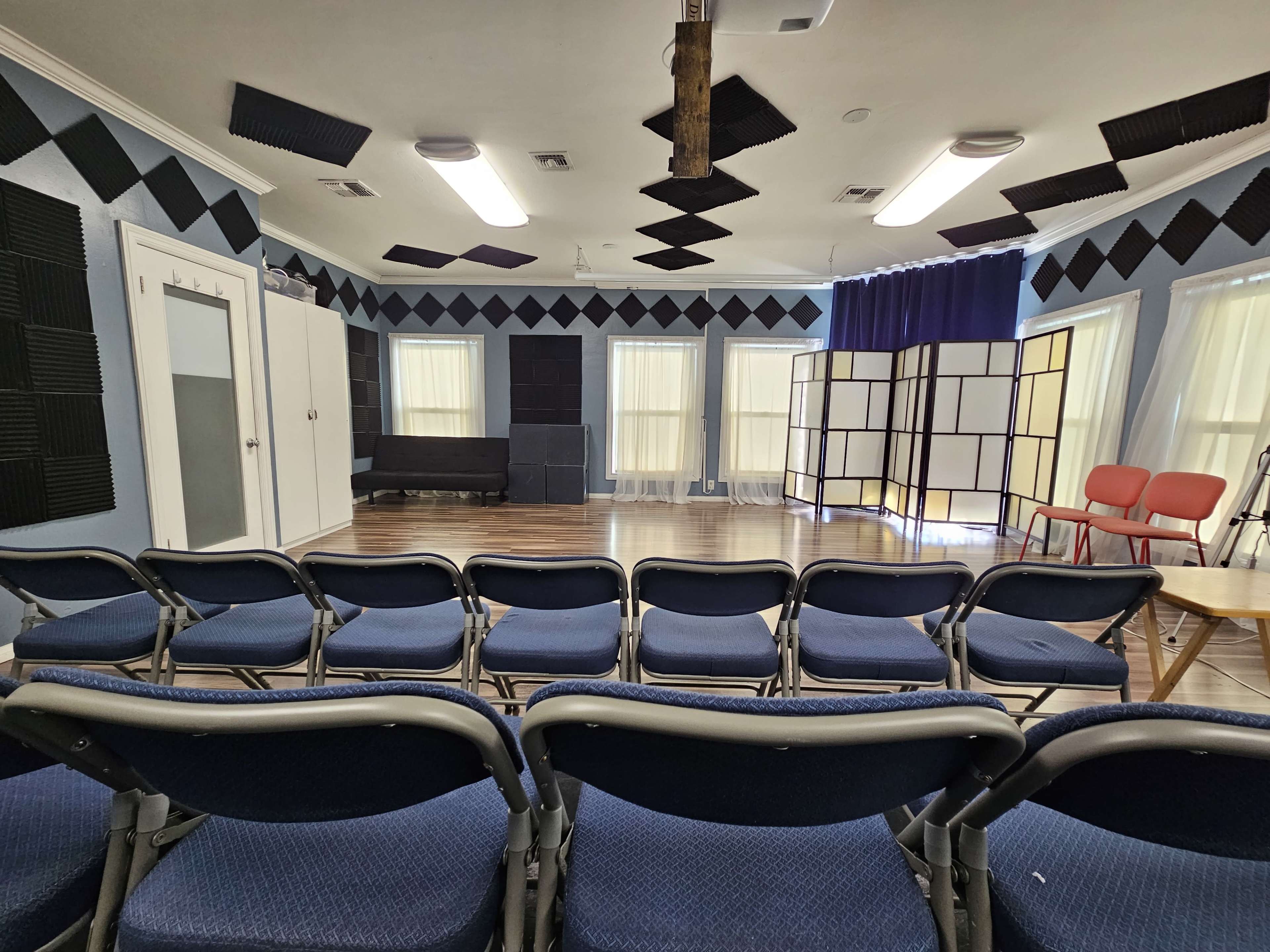The image shows a room set up for an event, featuring rows of blue chairs facing a stage area with soundproofing panels on the walls and a folding screen in the background.