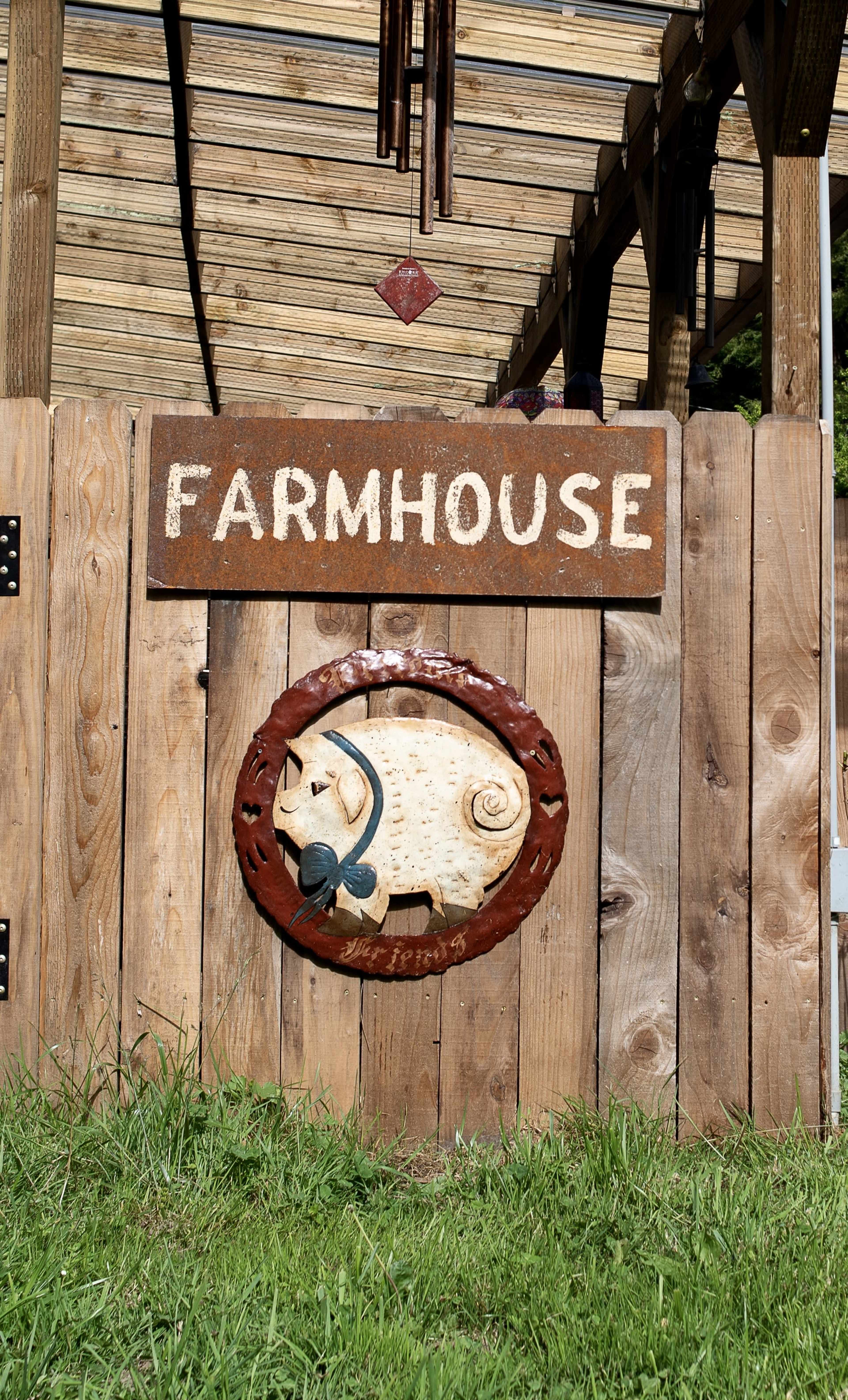 A wooden gate with a rusted sign reading "FARMHOUSE" and a decorative pig emblem is shown in a grassy area.