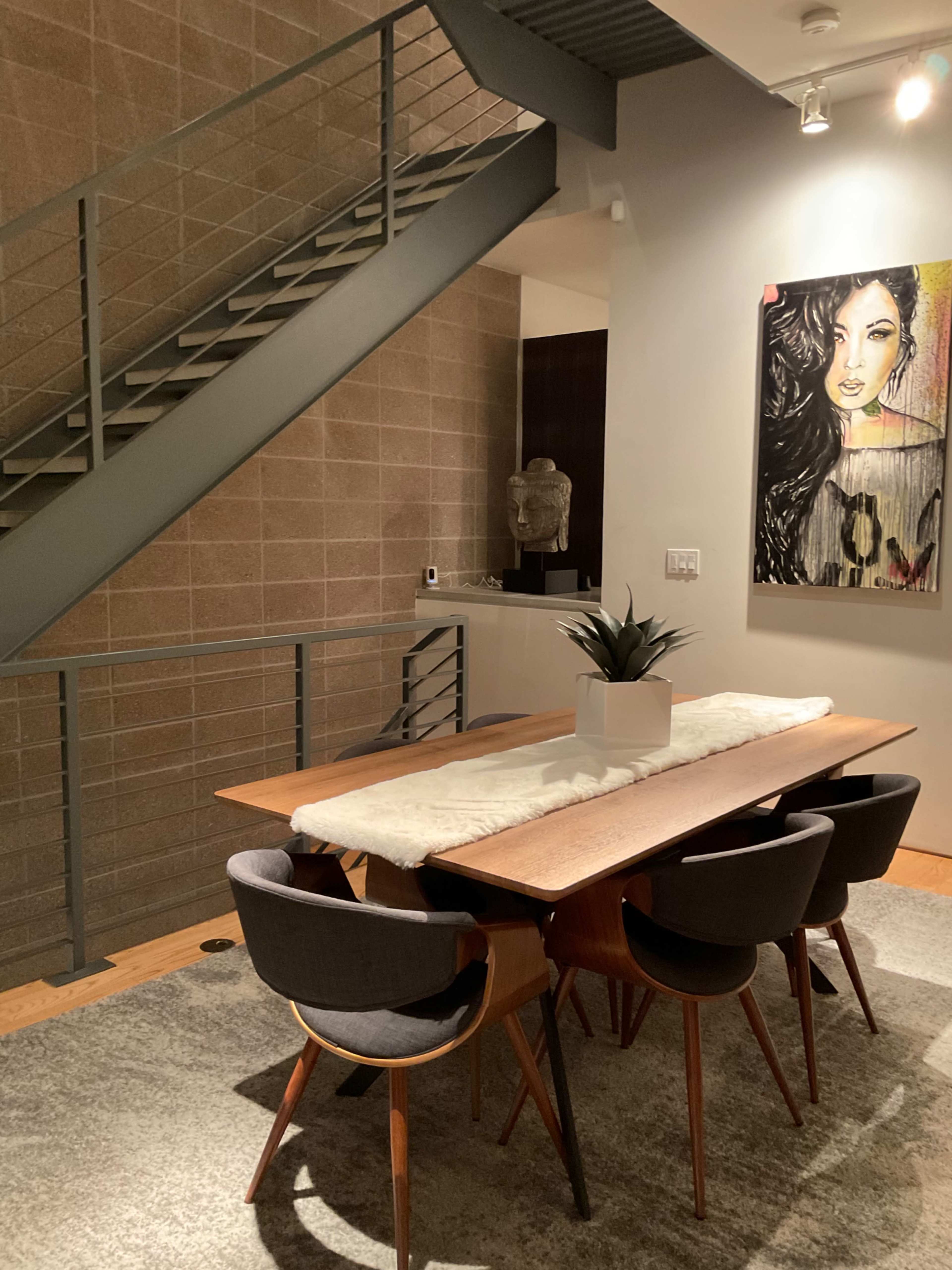 A dining area features a wooden table with upholstered chairs, a decorative plant in the center, a large artwork on the wall, and staircase access in the background.