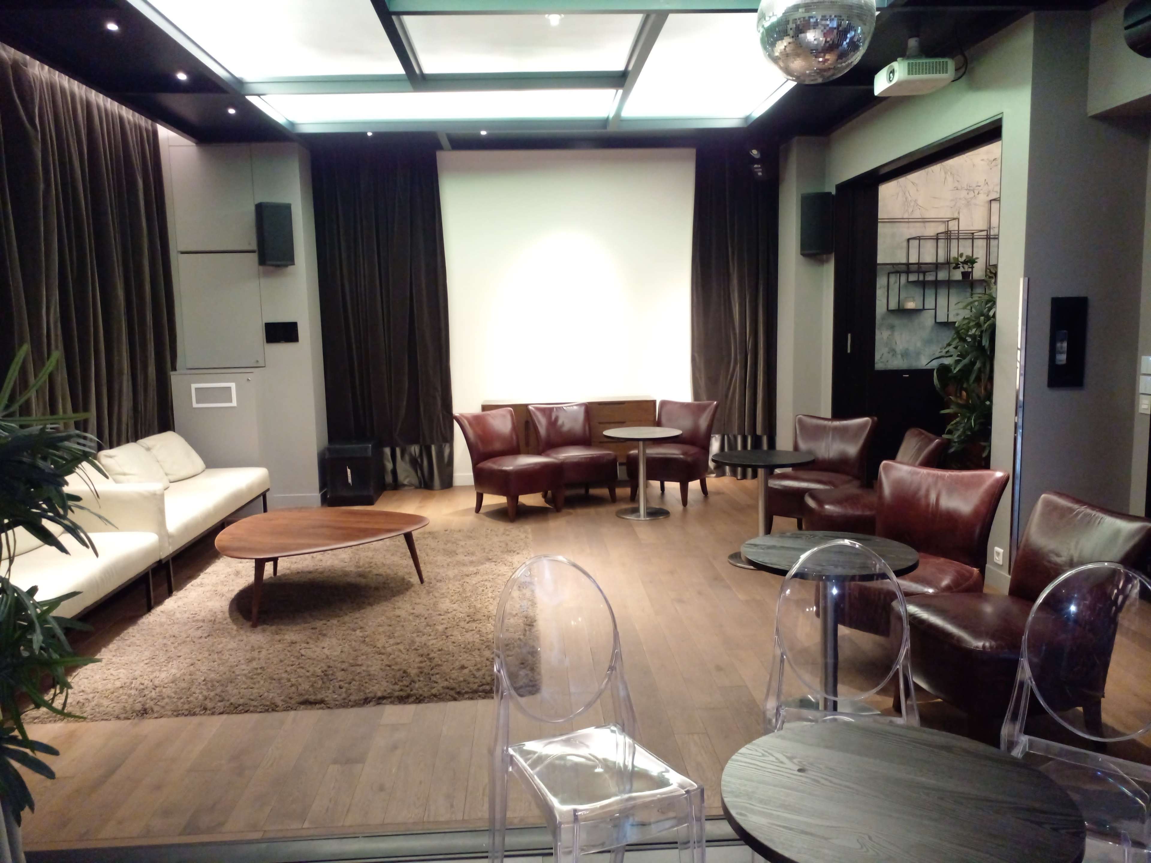 A modern lounge area with a white sofa, brown leather chairs, a wooden coffee table, and transparent chairs on a carpeted floor, illuminated by overhead lighting.