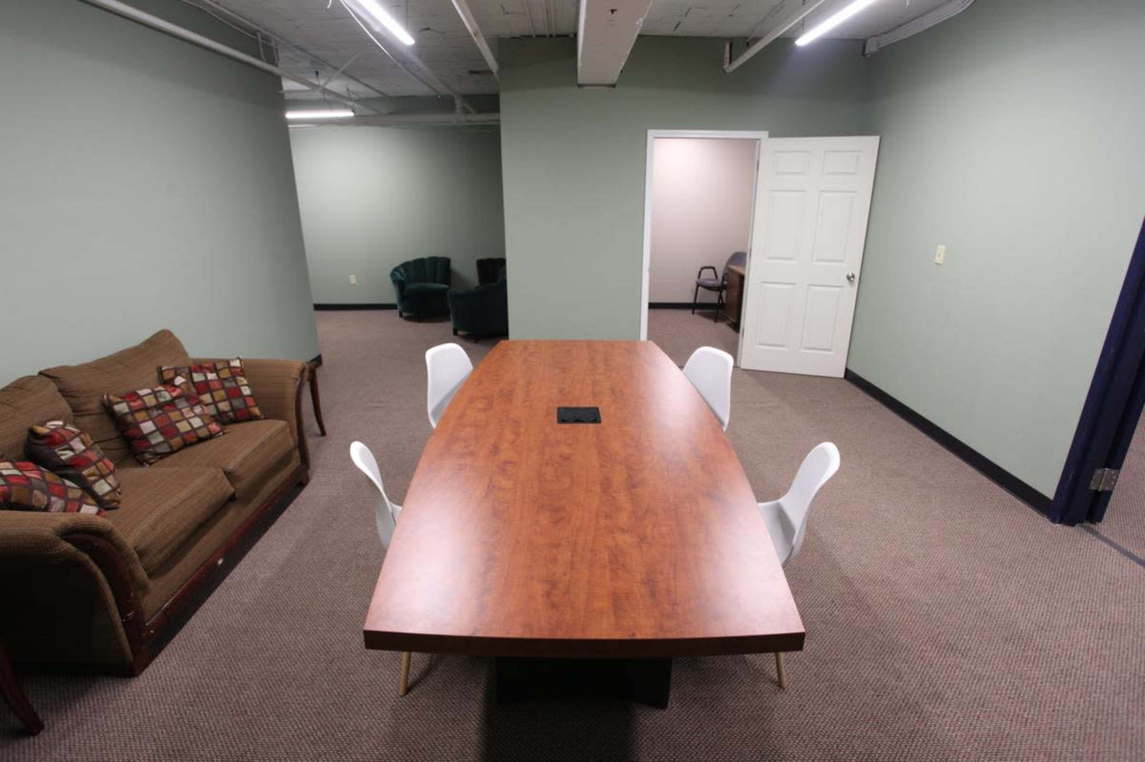 A conference room features a large wooden table surrounded by four white chairs, with a brown couch in the foreground and green walls.