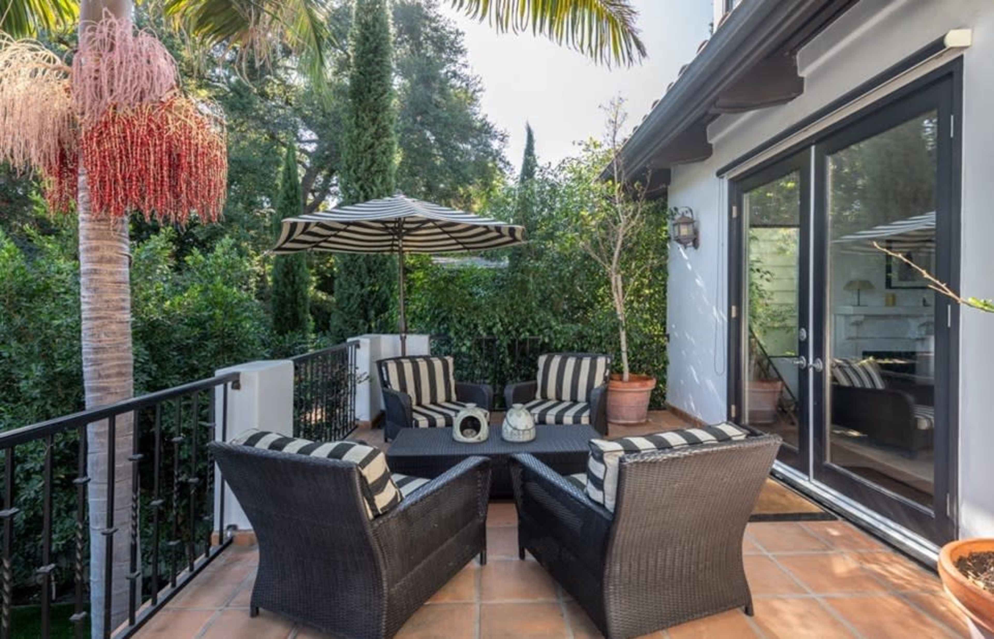 A patio features black wicker furniture with striped cushions, an umbrella, and potted plants surrounded by greenery.