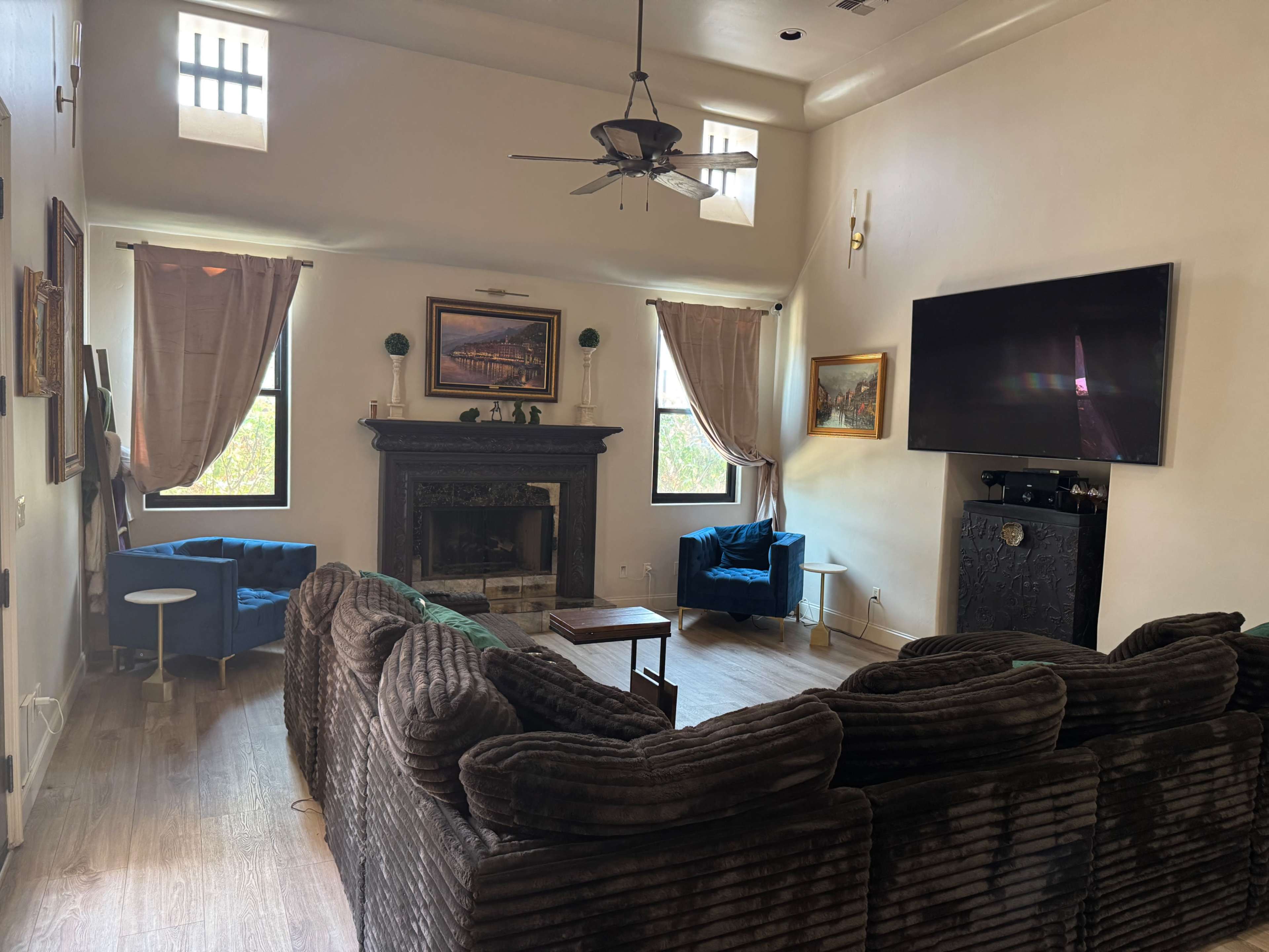 The image shows a spacious living room with a large, dark sofa, a fireplace, two blue armchairs, and a mounted TV on the wall.