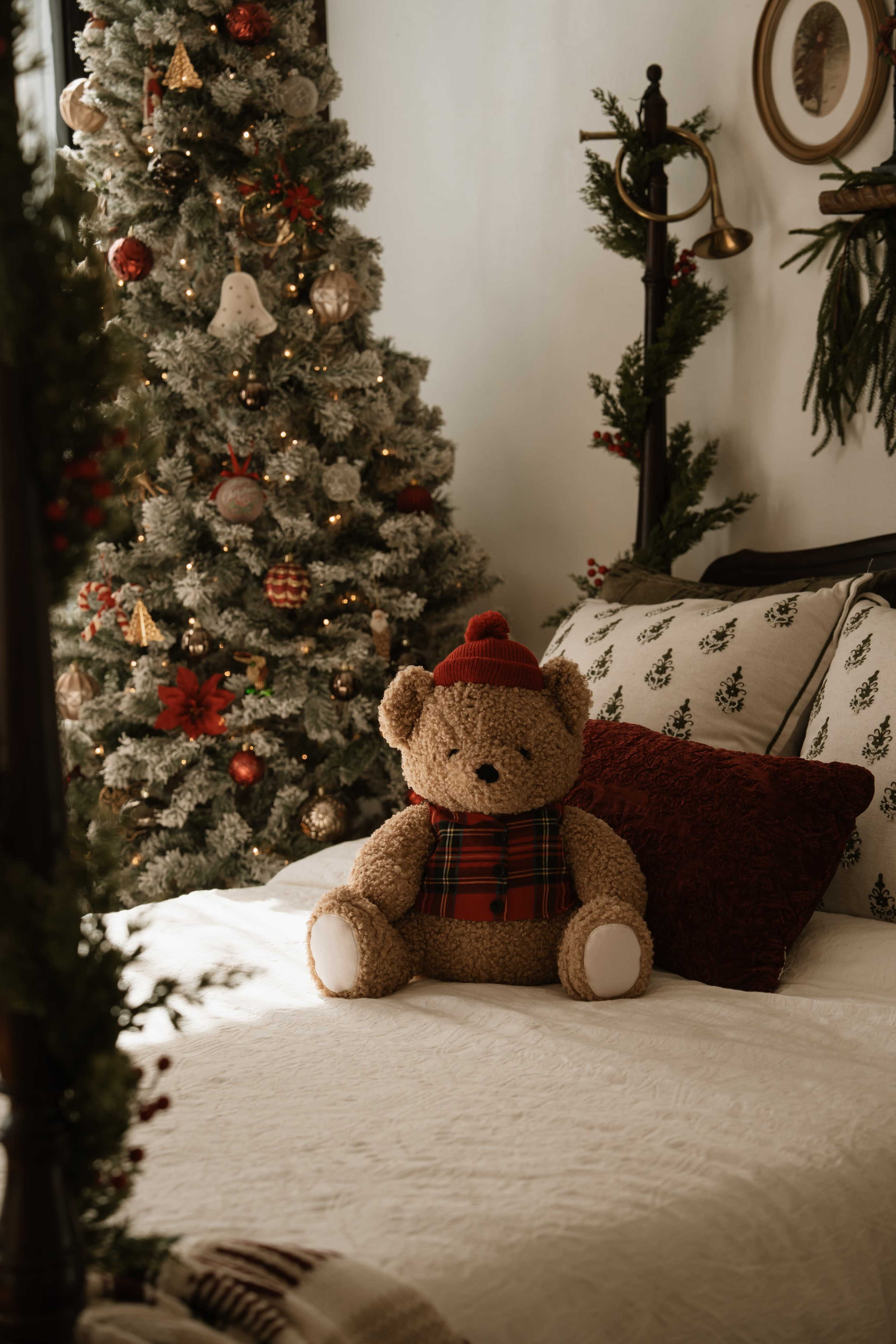 A large plush teddy bear dressed in a red hat and plaid scarf sits on a bed in a room decorated for the holidays, featuring a Christmas tree adorned with lights and ornaments in the background.
