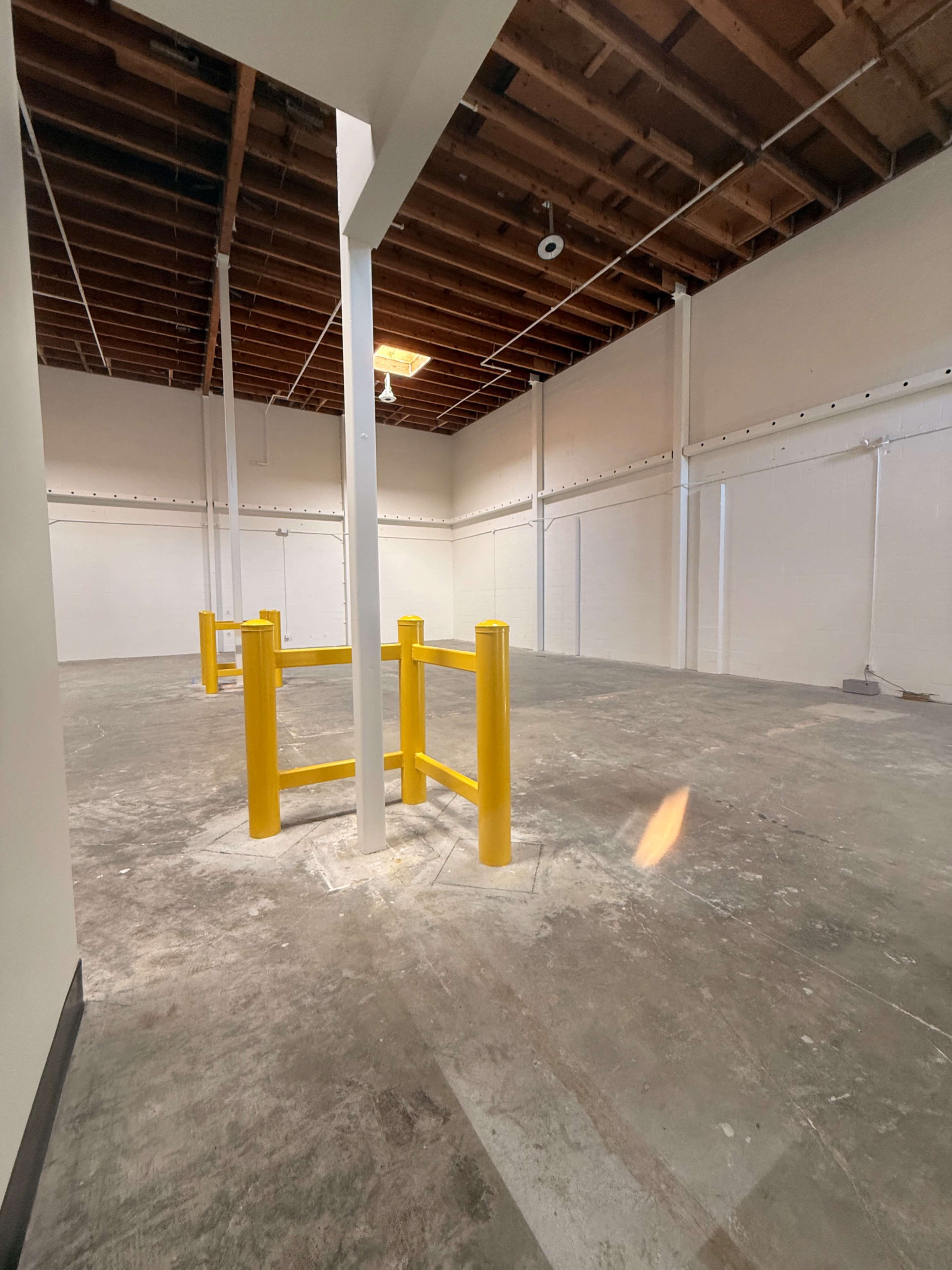 The image shows an empty industrial space with a concrete floor, white walls, and yellow safety bollards near the center.
