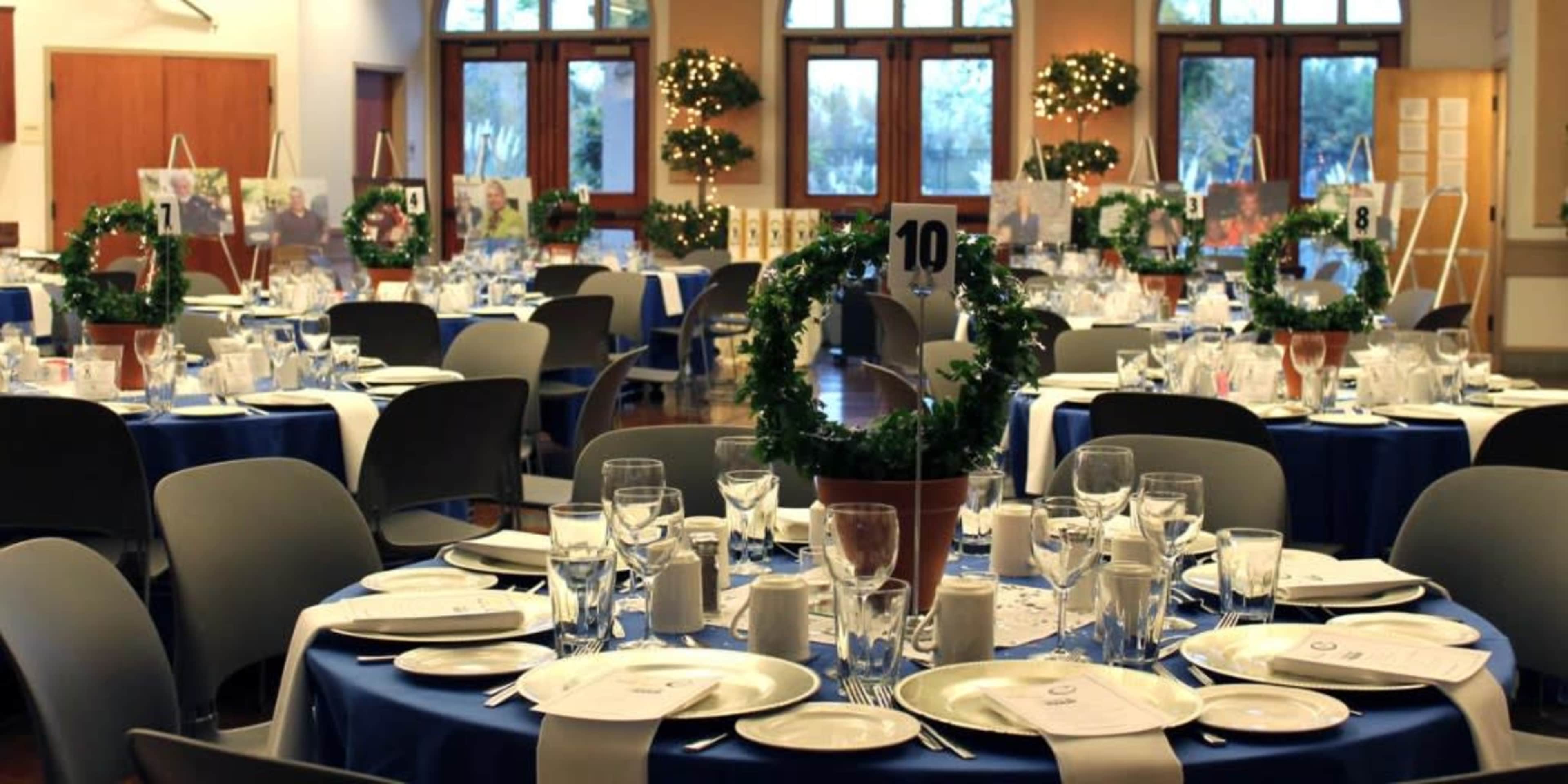 A banquet hall arranged with round tables set for dining, each featuring blue tablecloths, plates, glassware, and decorative centerpieces.