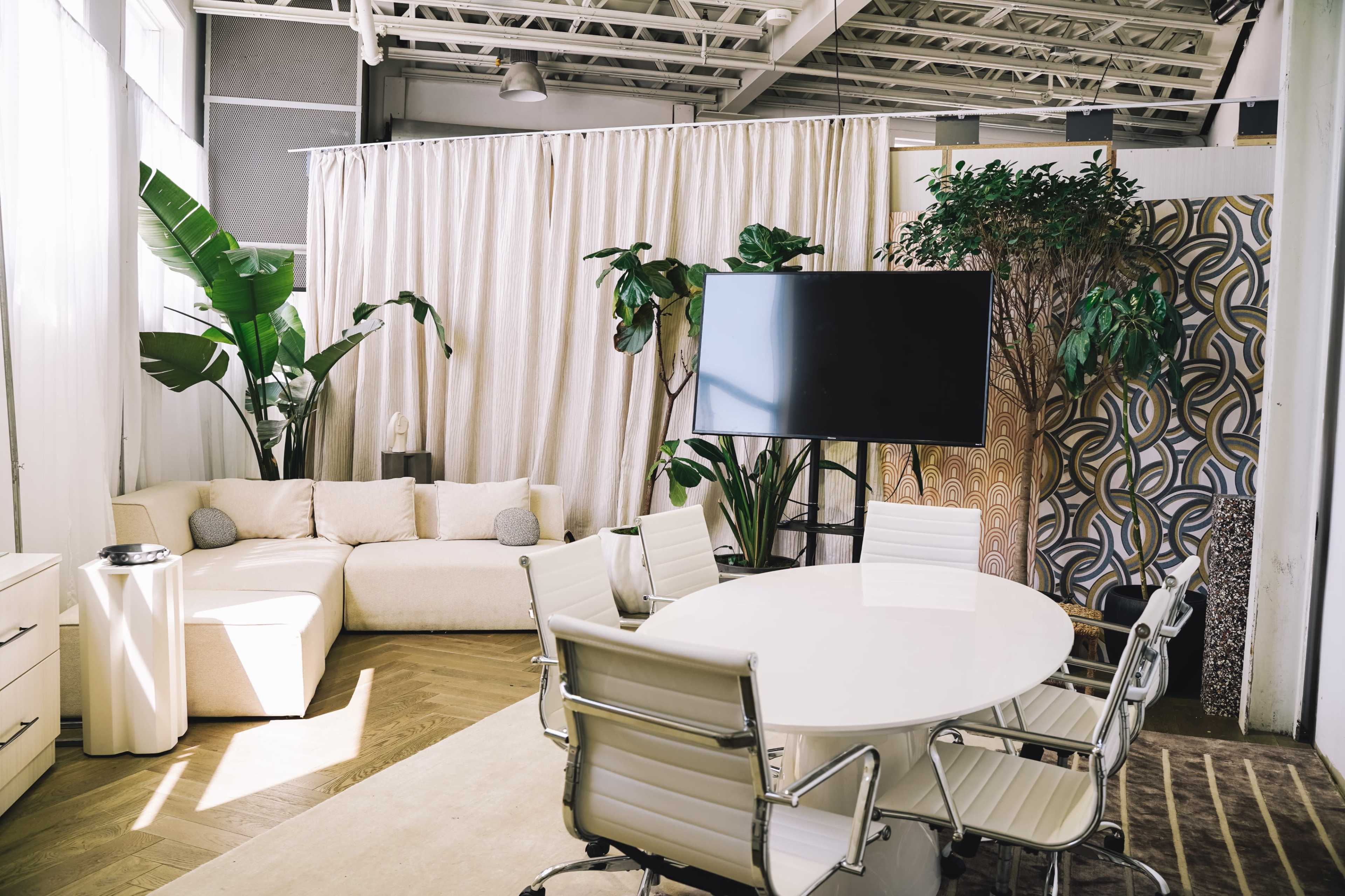 A modern meeting room featuring a round table surrounded by white chairs, a large television screen, a sectional sofa, and indoor plants.