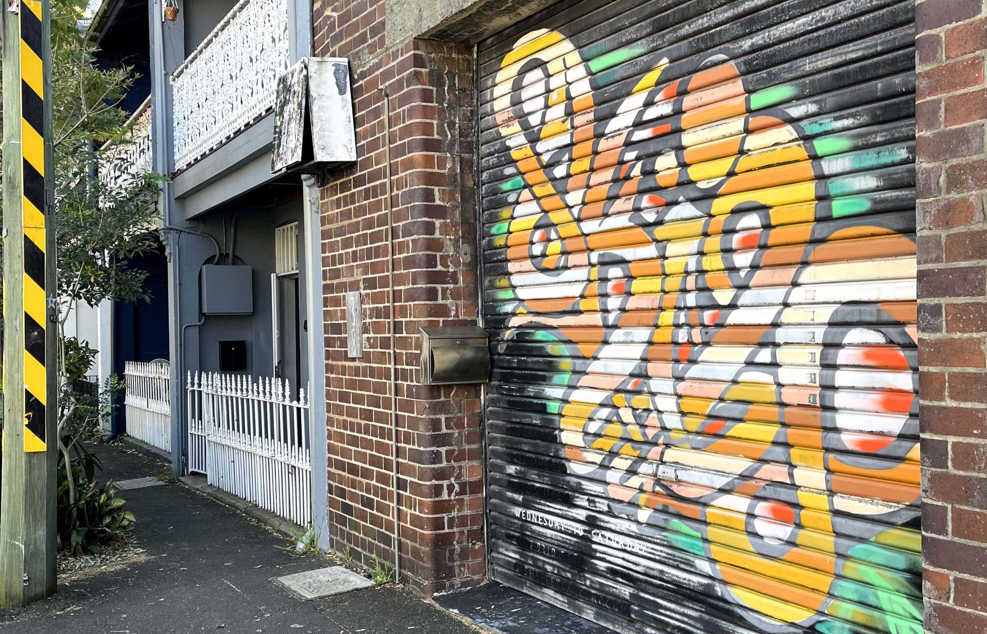 A colorful graffiti mural covers a rolled-down garage door next to a narrow pathway lined with a white picket fence.
