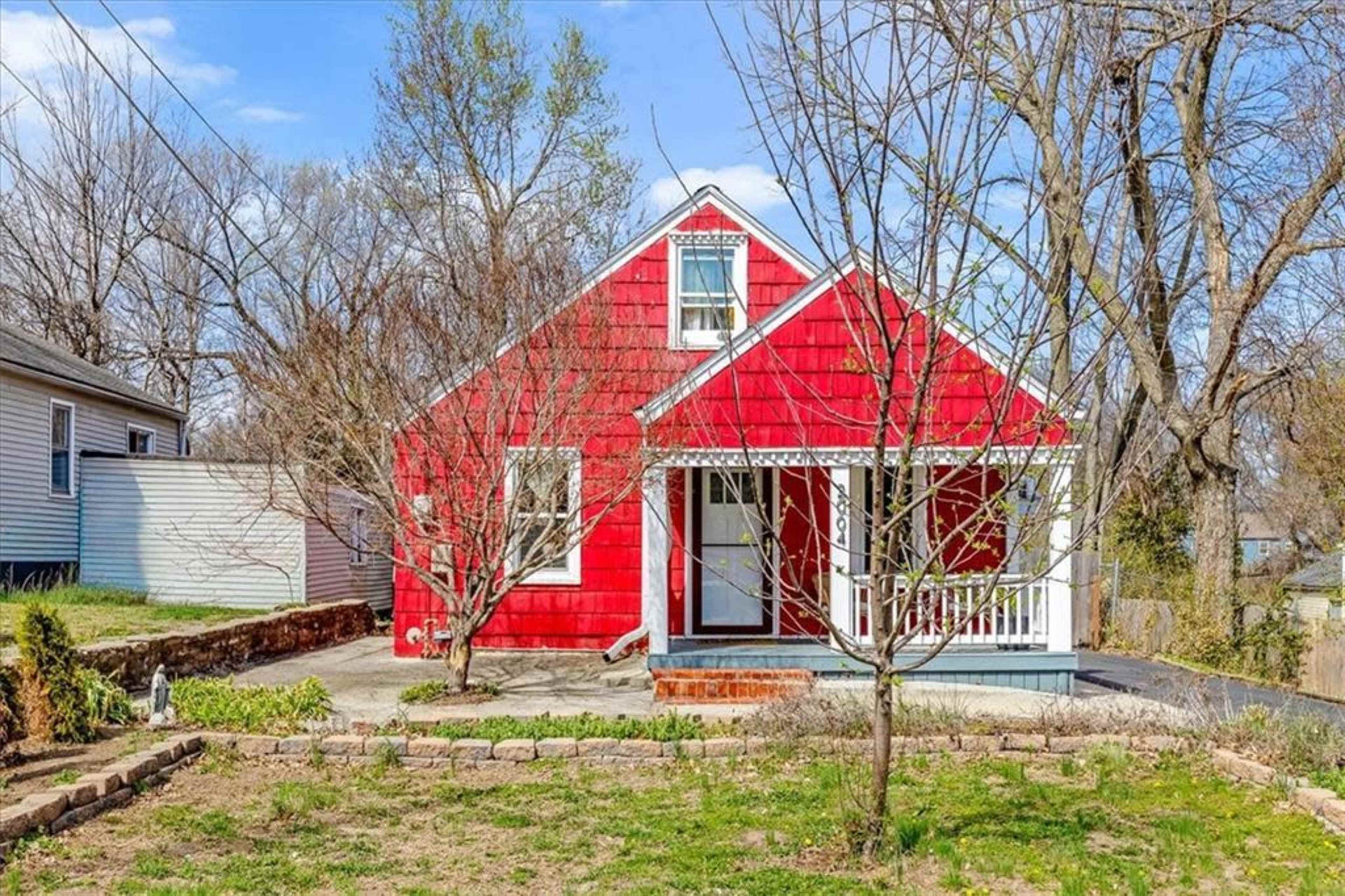 The image shows a red house with a gabled roof, a front porch, and a bare yard bordered by trees and shrubs.