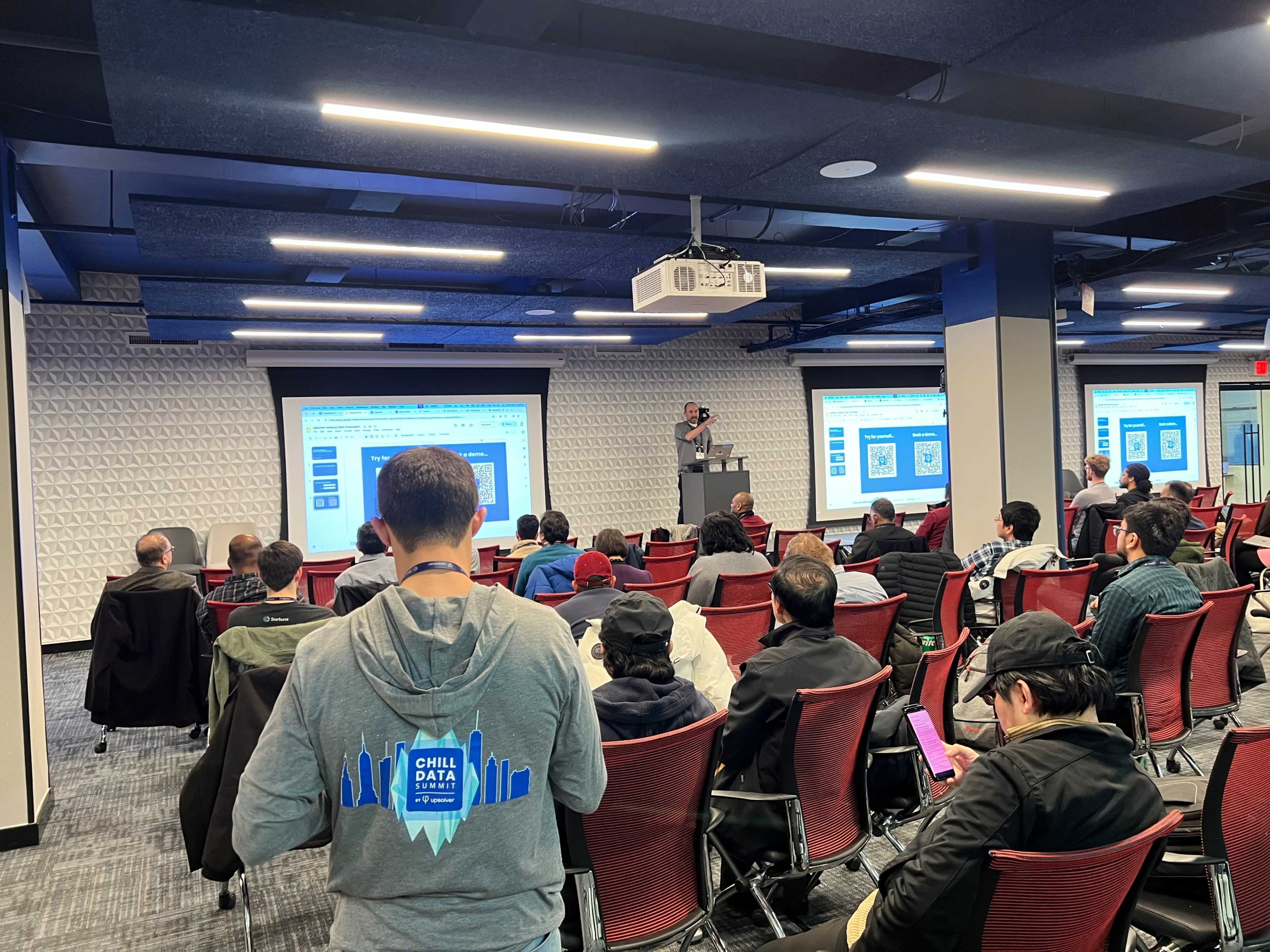 A speaker presents to an audience in a modern conference room equipped with large screens and rows of chairs.