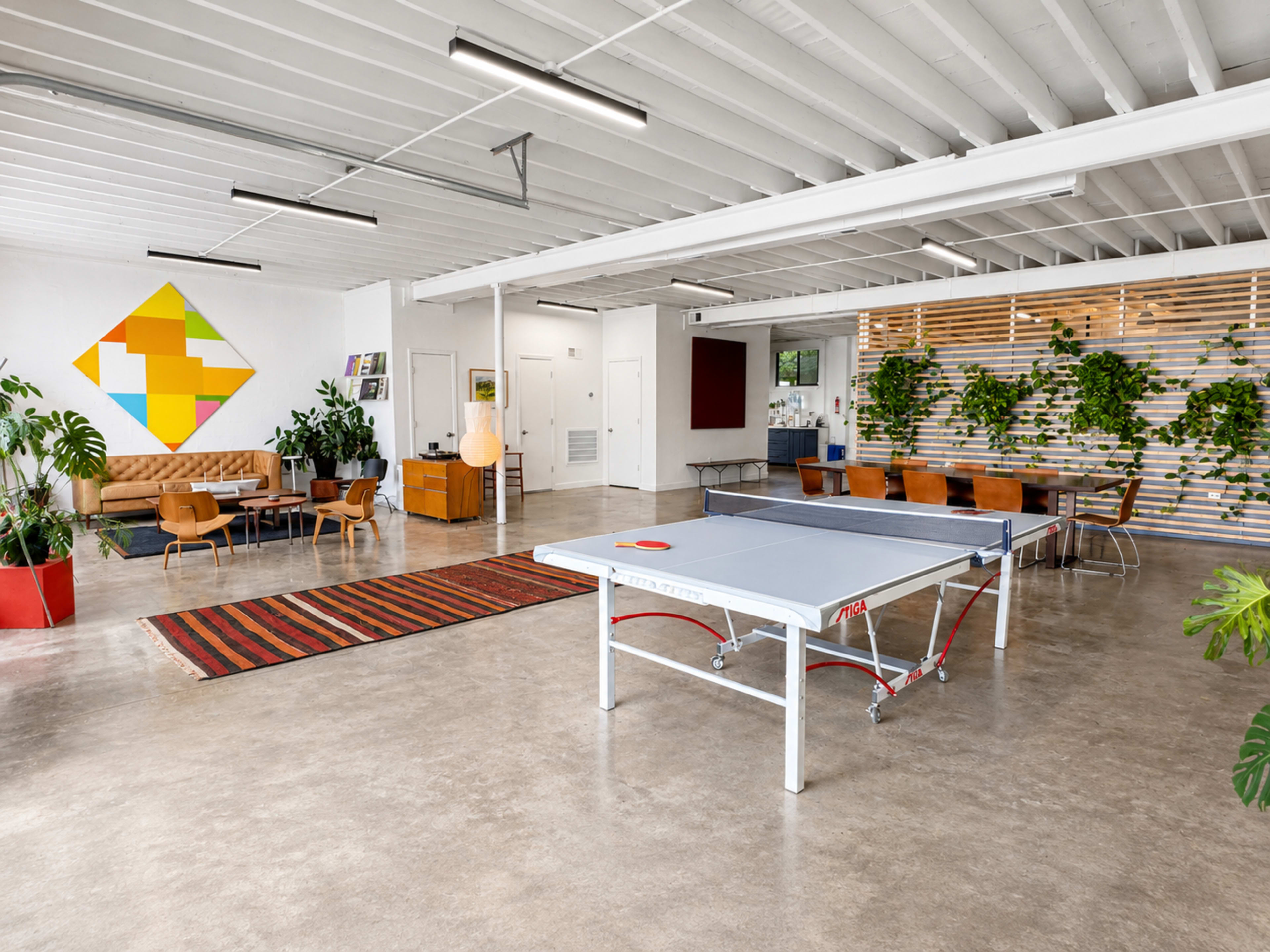 The image shows a spacious, modern office interior with a ping pong table in the center, surrounded by seating areas, decorative plants, and a colorful wall design.