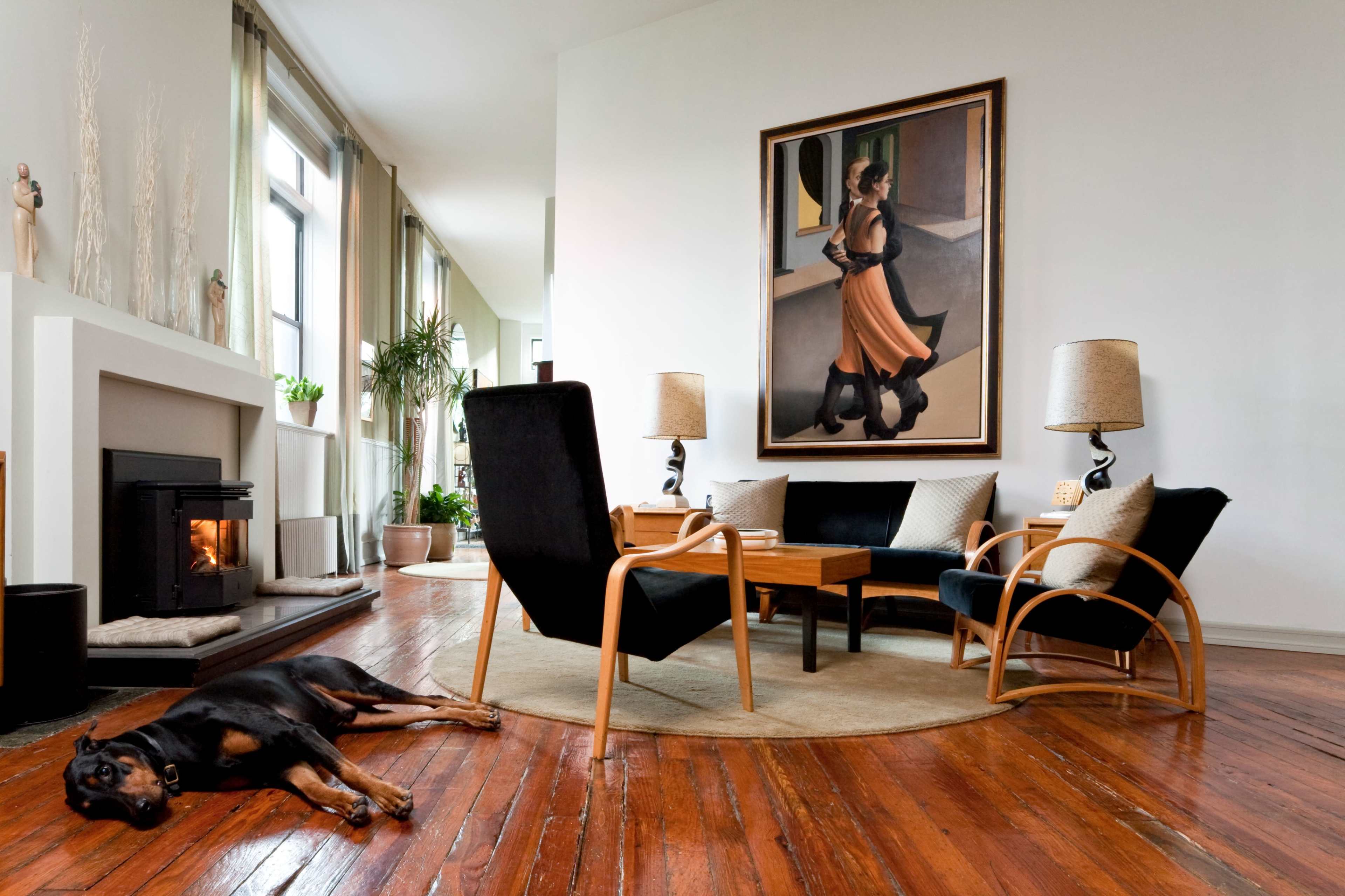 A spacious living room features a painting of a woman on the wall, a coffee table surrounded by two armchairs, and a dog resting on the wooden floor.