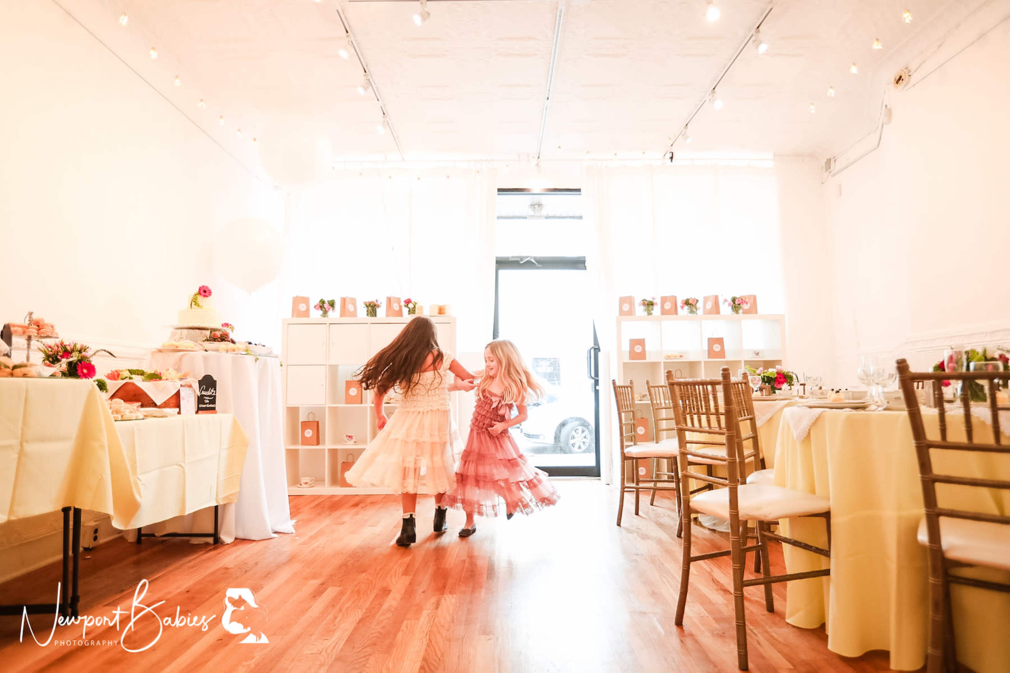 Two young girls in pink dresses dance playfully in a softly lit room set for a celebration, with tables arranged and decorations in the background.