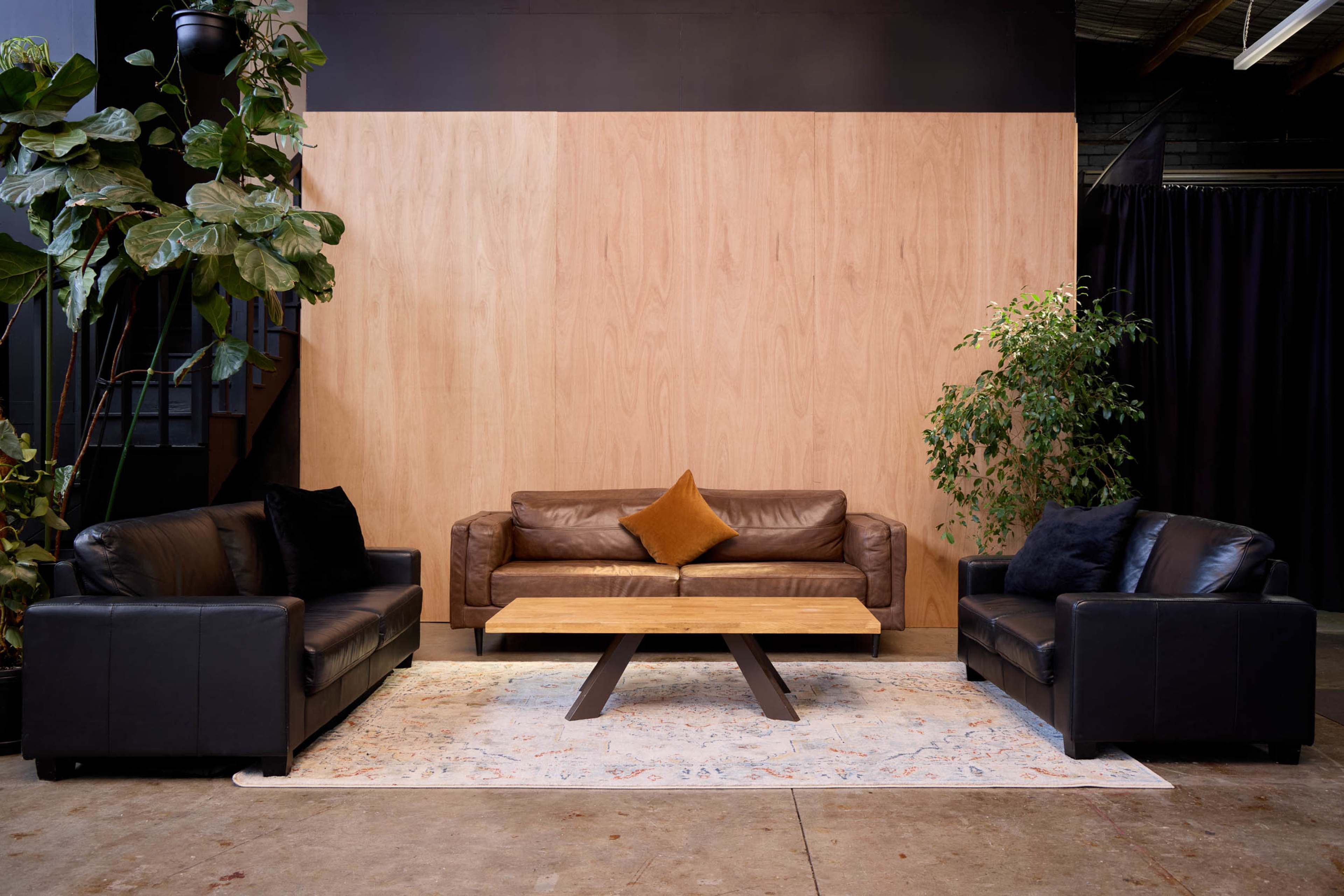 A modern living room features two black leather sofas and a tan leather couch around a wooden coffee table, set against a wooden panel backdrop and surrounded by indoor plants.