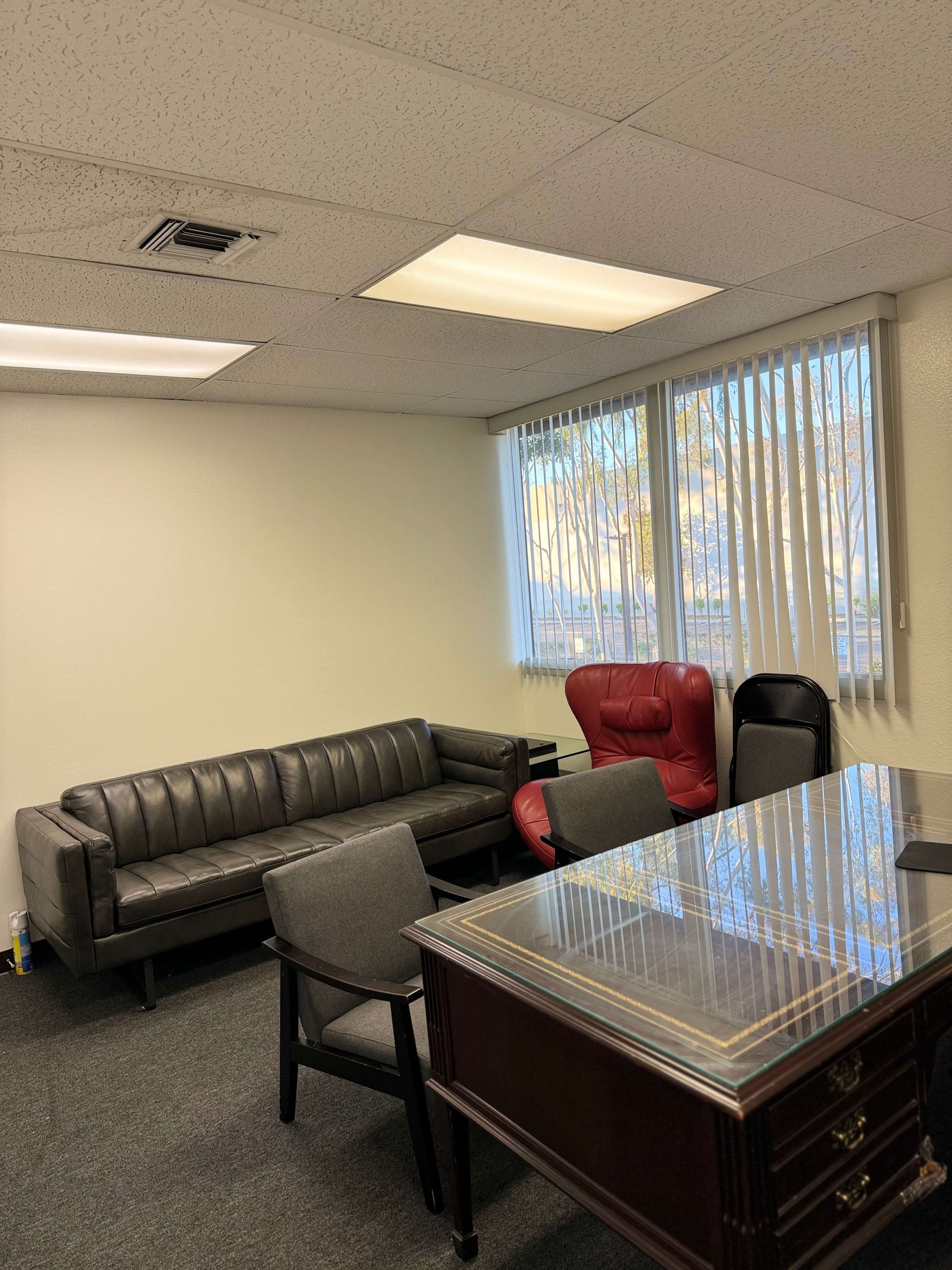 An office space with a black leather couch, a red armchair, several gray chairs, and a wooden desk with a glass top, illuminated by natural light from a window.