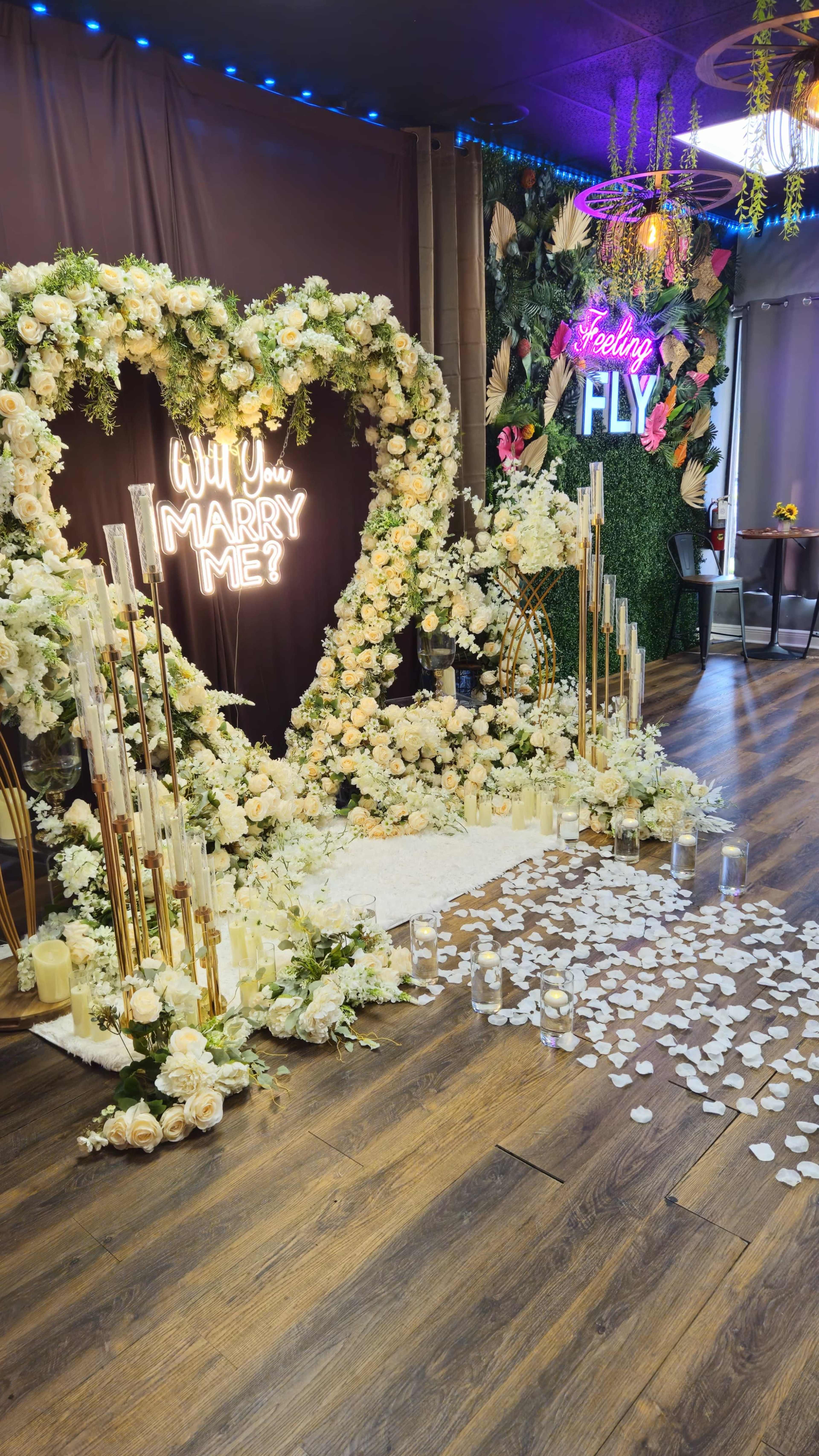 The image shows a wedding proposal setup featuring a heart-shaped floral arrangement, a "Will You Marry Me?" sign in neon lights, a pathway of rose petals, and candles lining the ground.