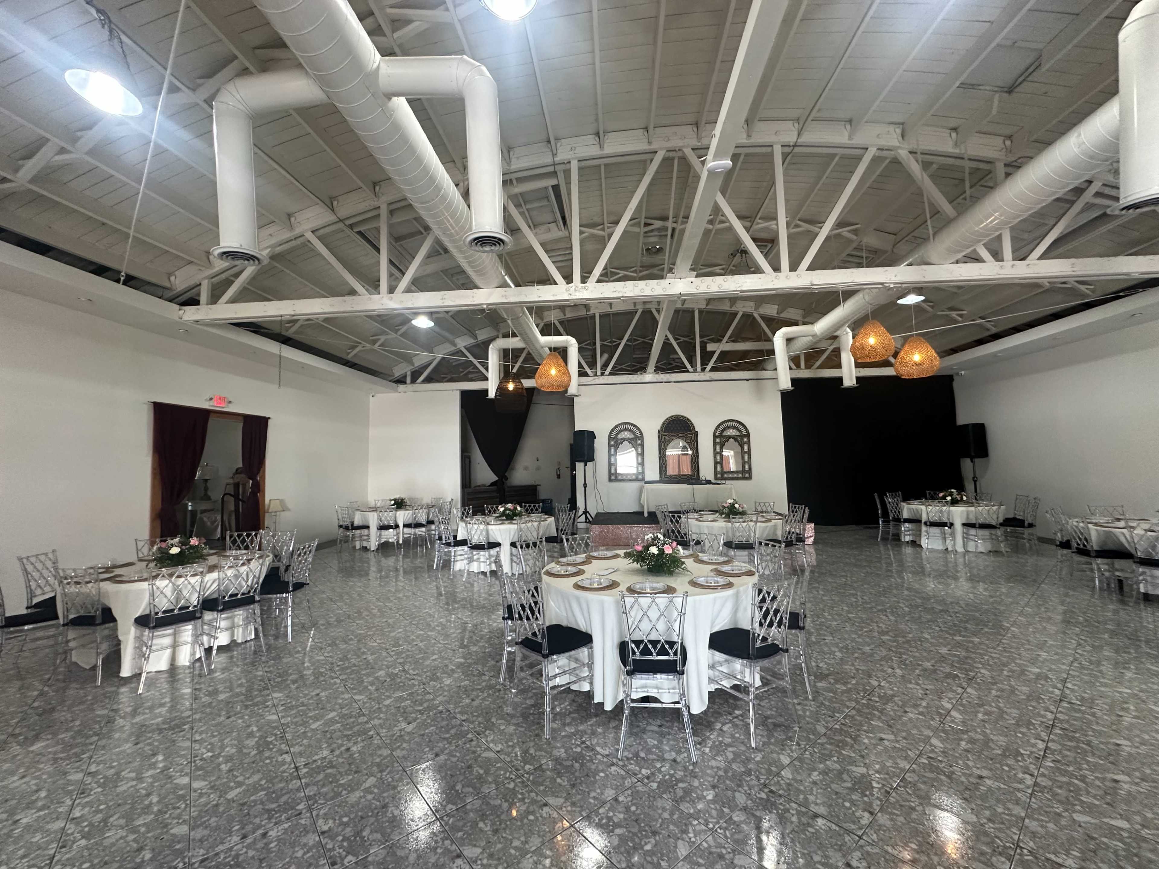 The image shows a spacious event venue with round tables set for a gathering, featuring a polished floor and exposed ceiling beams.