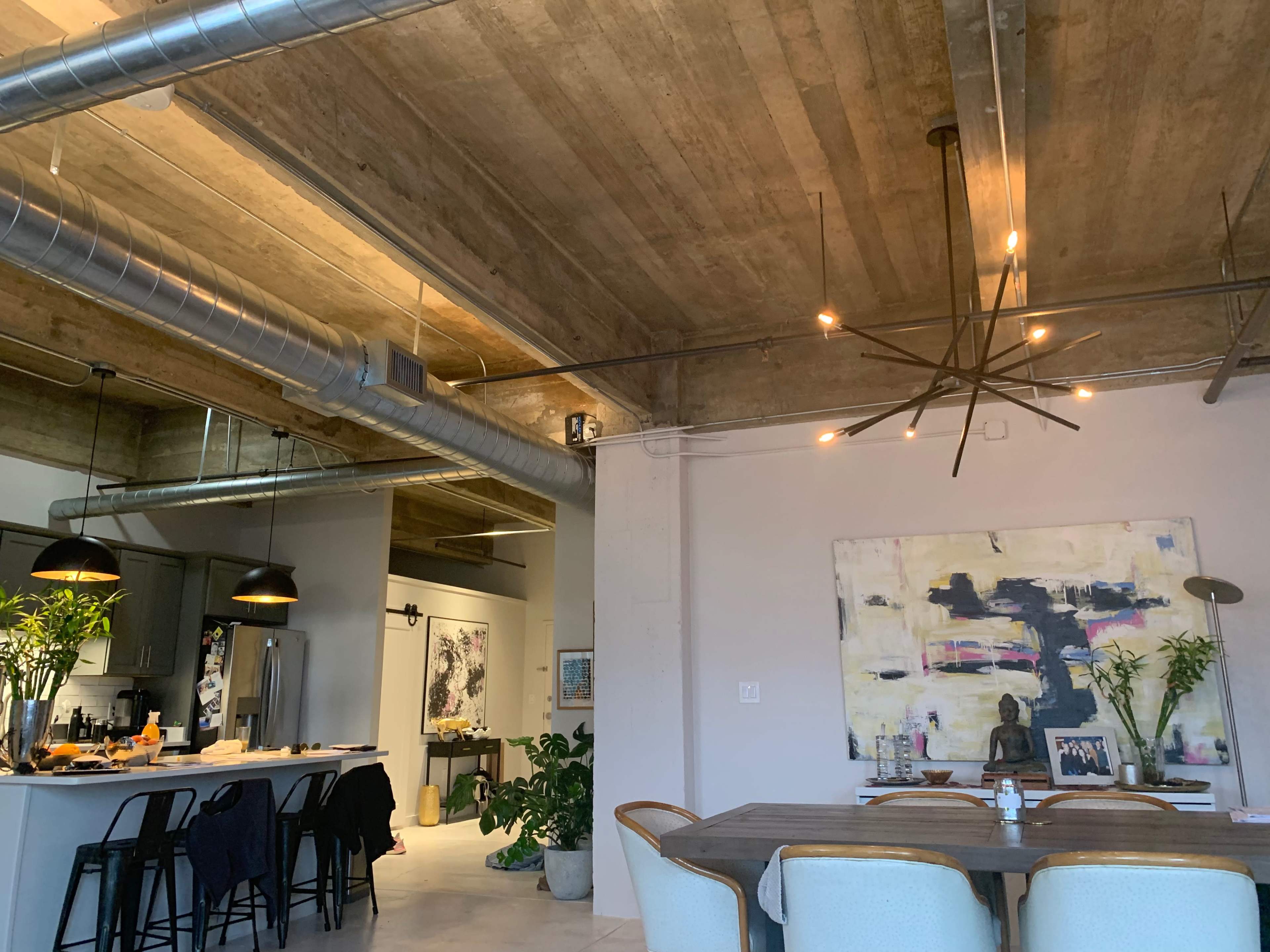 The image shows a modern open-concept loft with exposed concrete ceilings, a dining table, and a spacious kitchen area featuring industrial-style lighting fixtures.