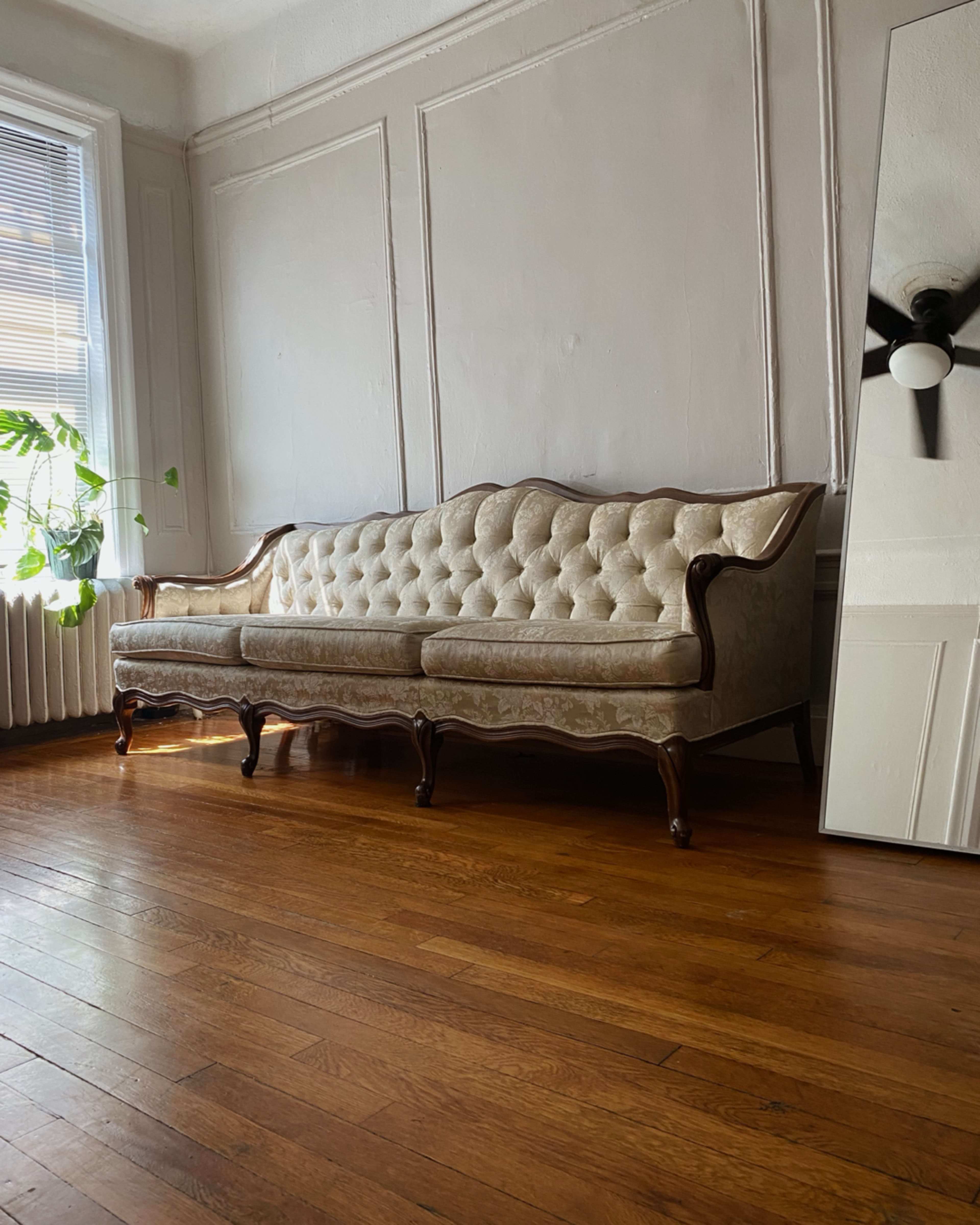 A vintage tufted couch with wooden legs is positioned against a wall in a room with hardwood floors and a large mirror nearby.
