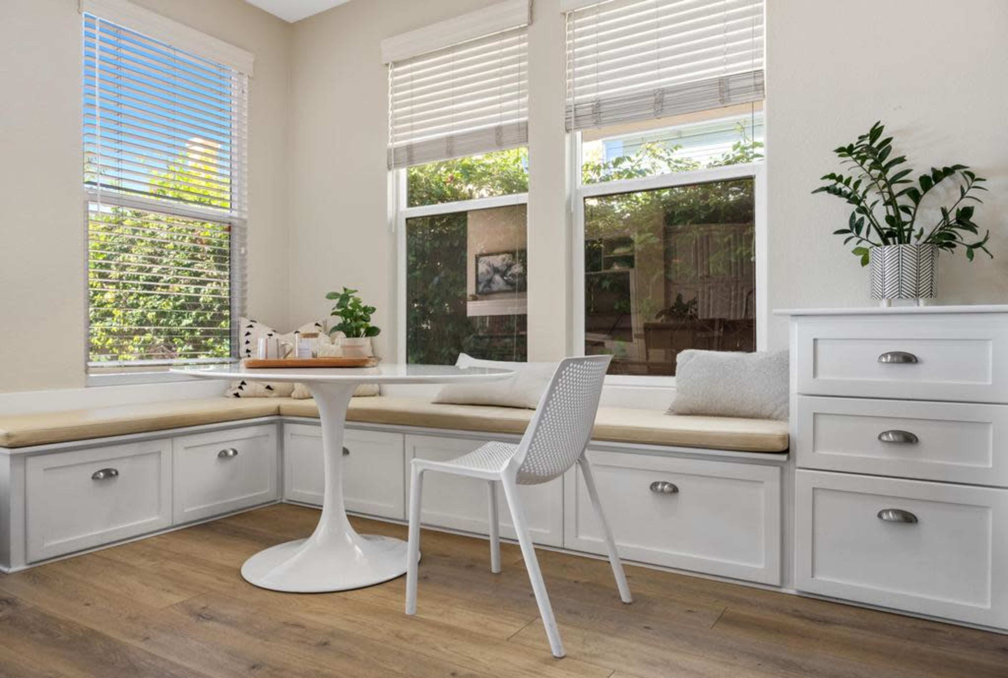 A bright corner nook features a built-in bench with storage, a small round table, a white chair, and large windows allowing natural light.