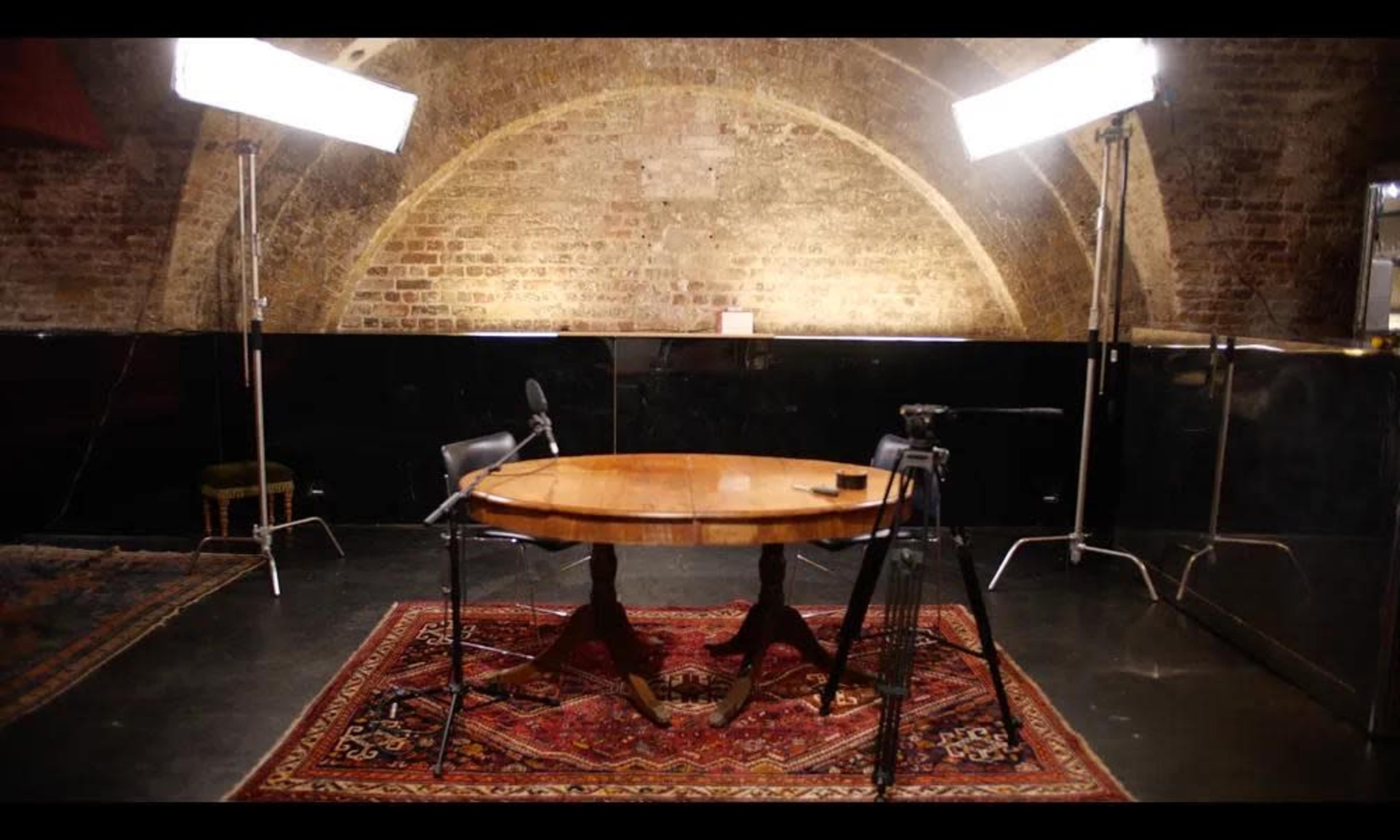 The image shows a dimly lit studio with a wooden table and two chairs, flanked by two studio lights and cameras.