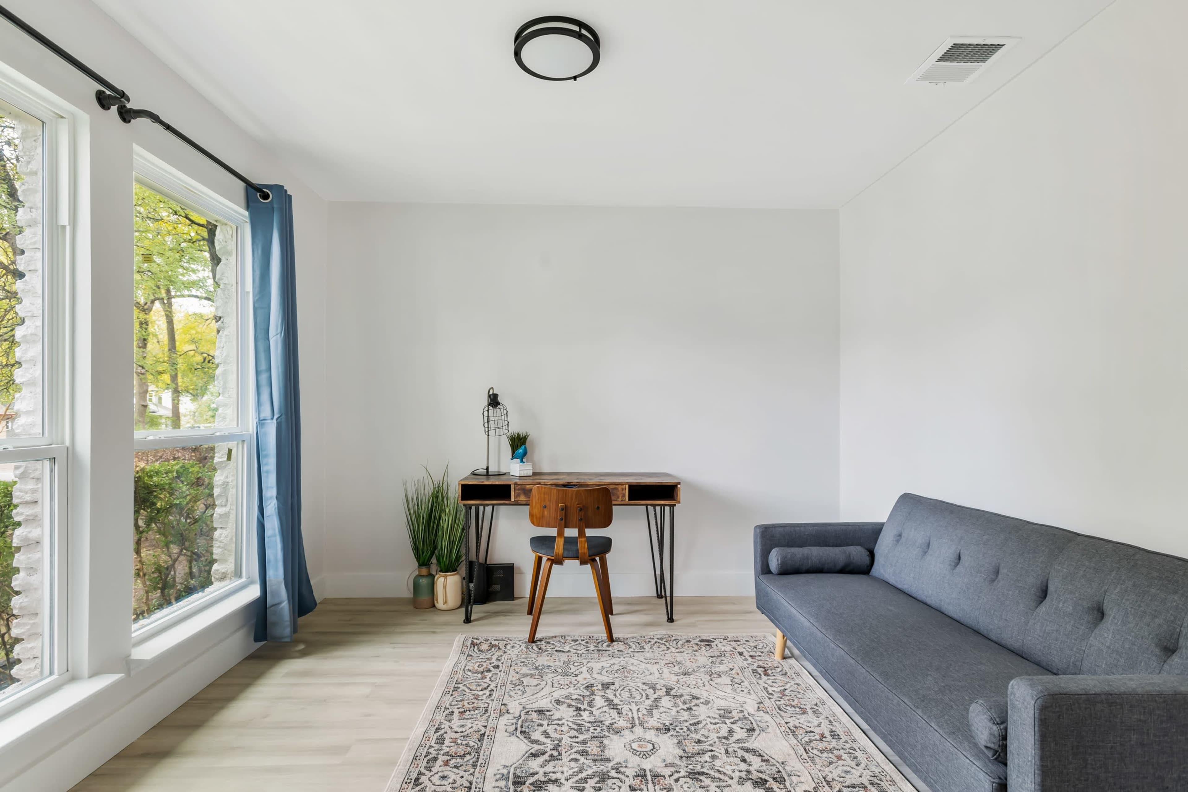 The room features a gray sofa, a wooden desk with a chair, a patterned rug, and large windows with light blue curtains.
