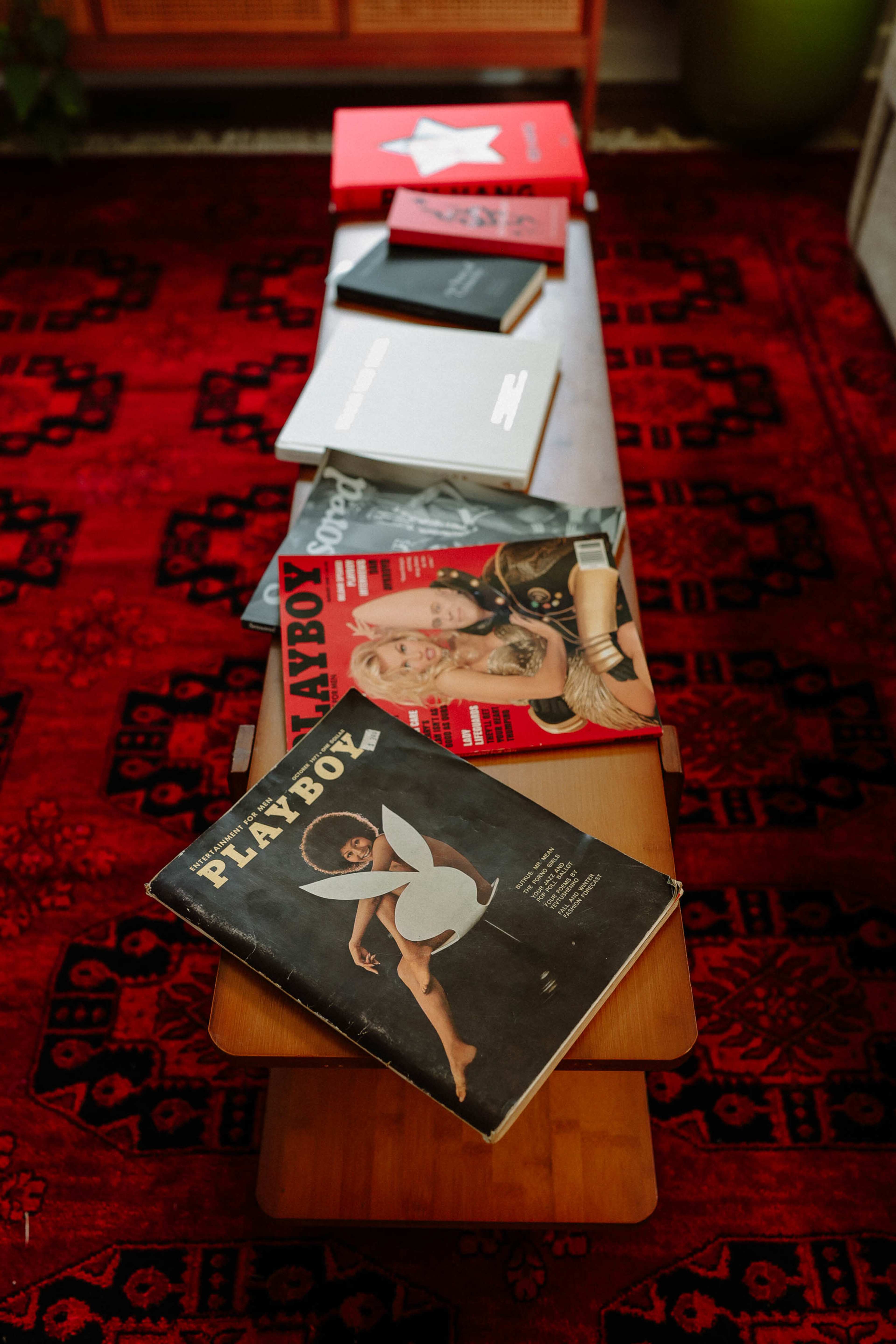 A wooden table displays a row of magazines, including several issues of Playboy, arranged on a patterned rug.