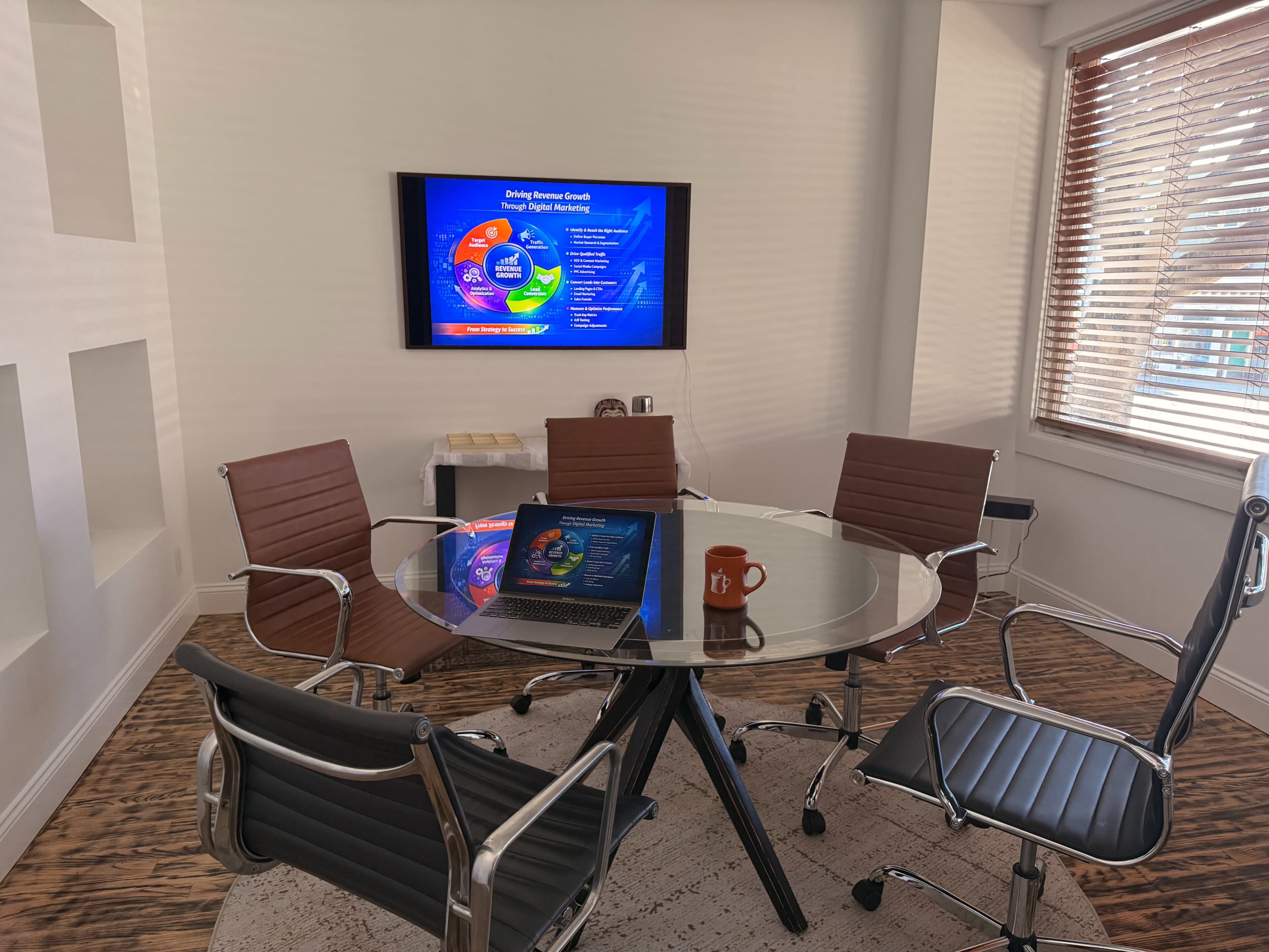 A modern conference room features a glass table surrounded by four leather chairs, with a laptop open and a presentation displayed on a wall-mounted screen.