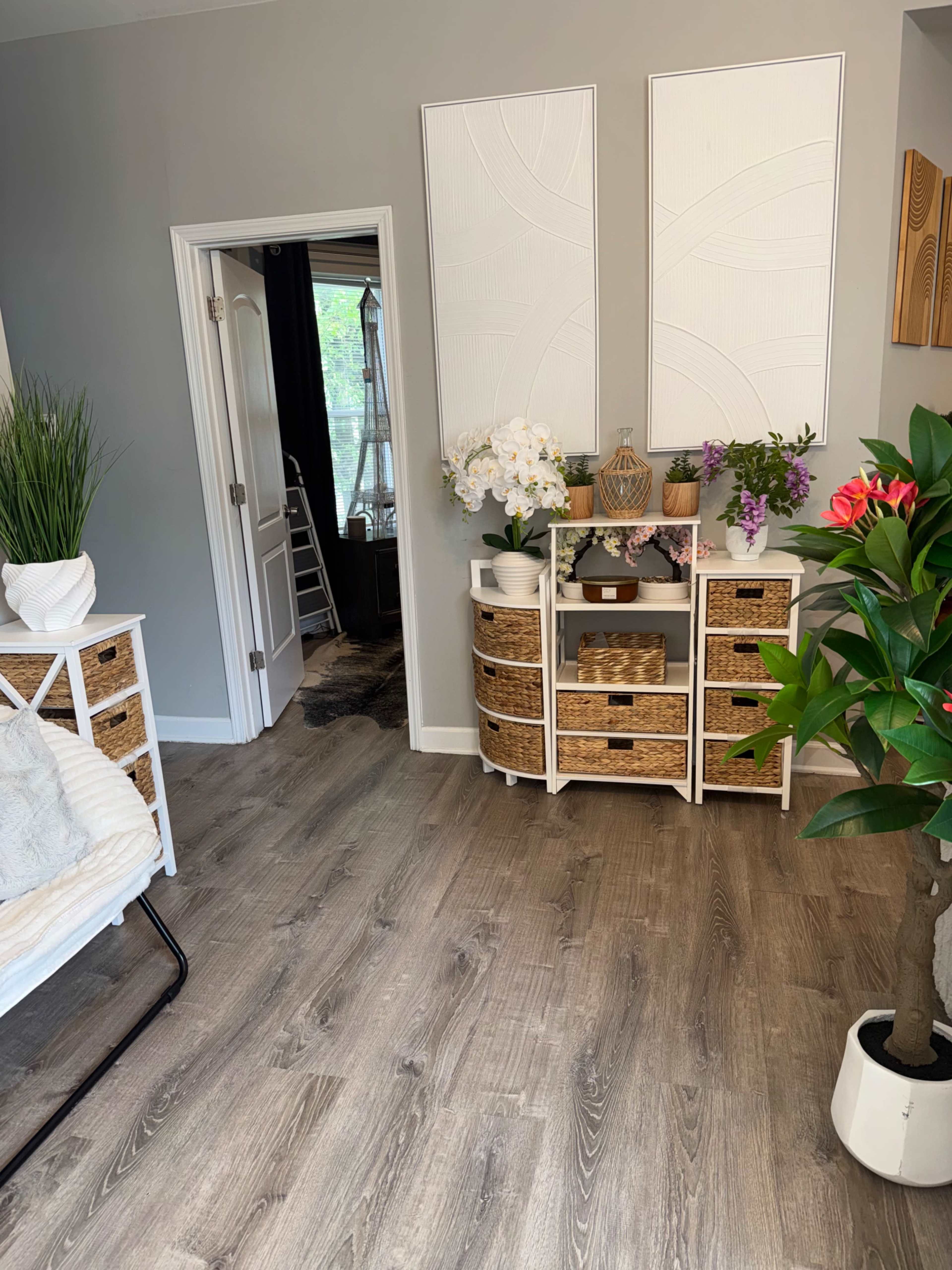 The image shows a cozy interior space featuring a light-colored floor, a white shelving unit with woven baskets, two decorative wall panels, and various potted plants.