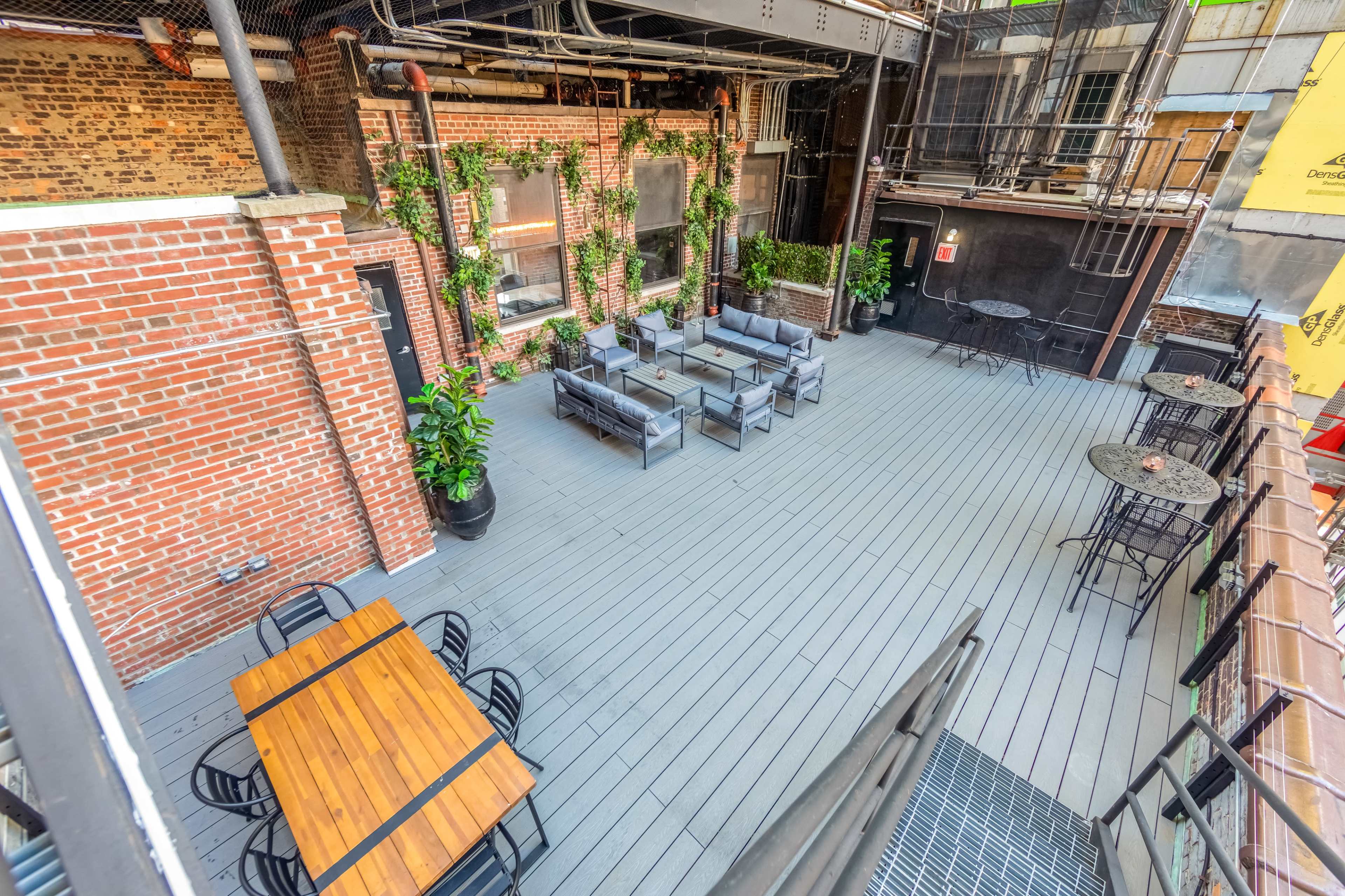A spacious outdoor patio area featuring wooden tables and chairs, surrounded by brick walls and greenery.