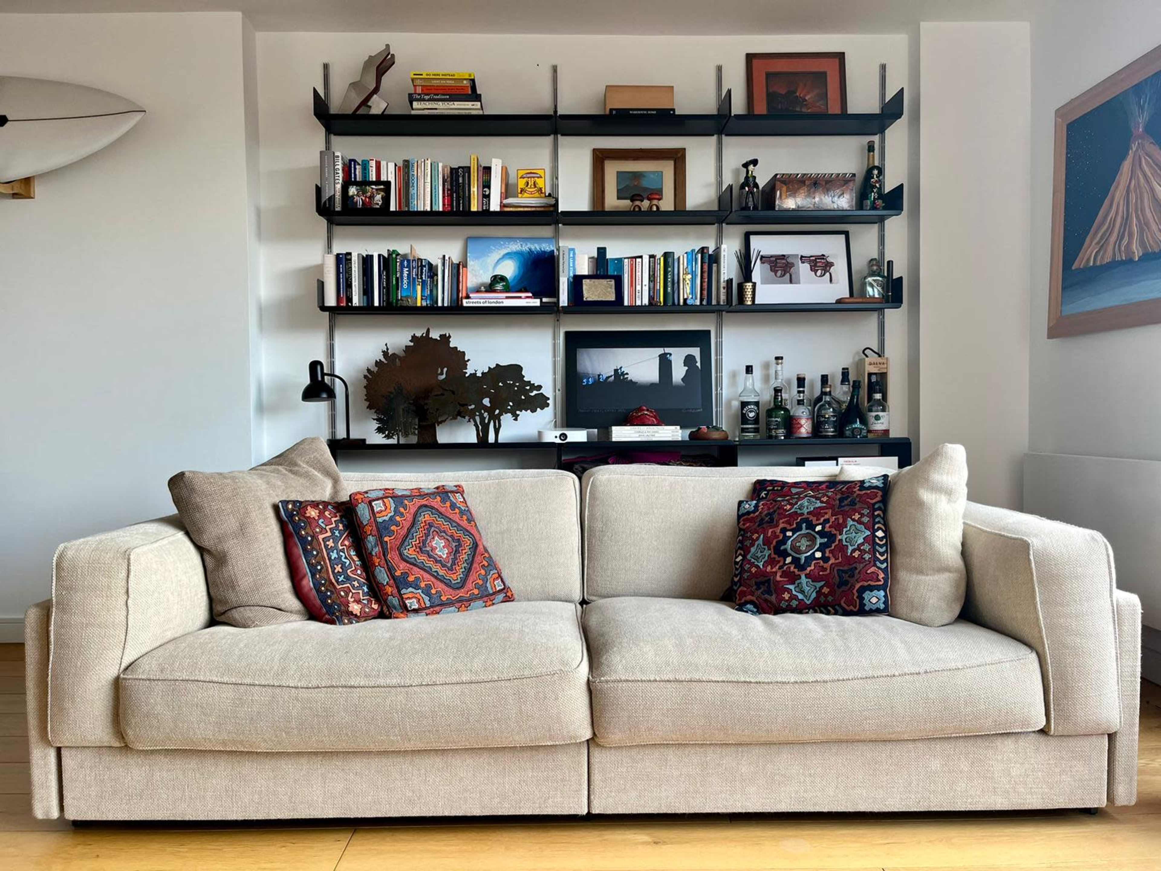 A beige couch with decorative pillows is positioned in front of a black bookshelf filled with books and various framed pictures.