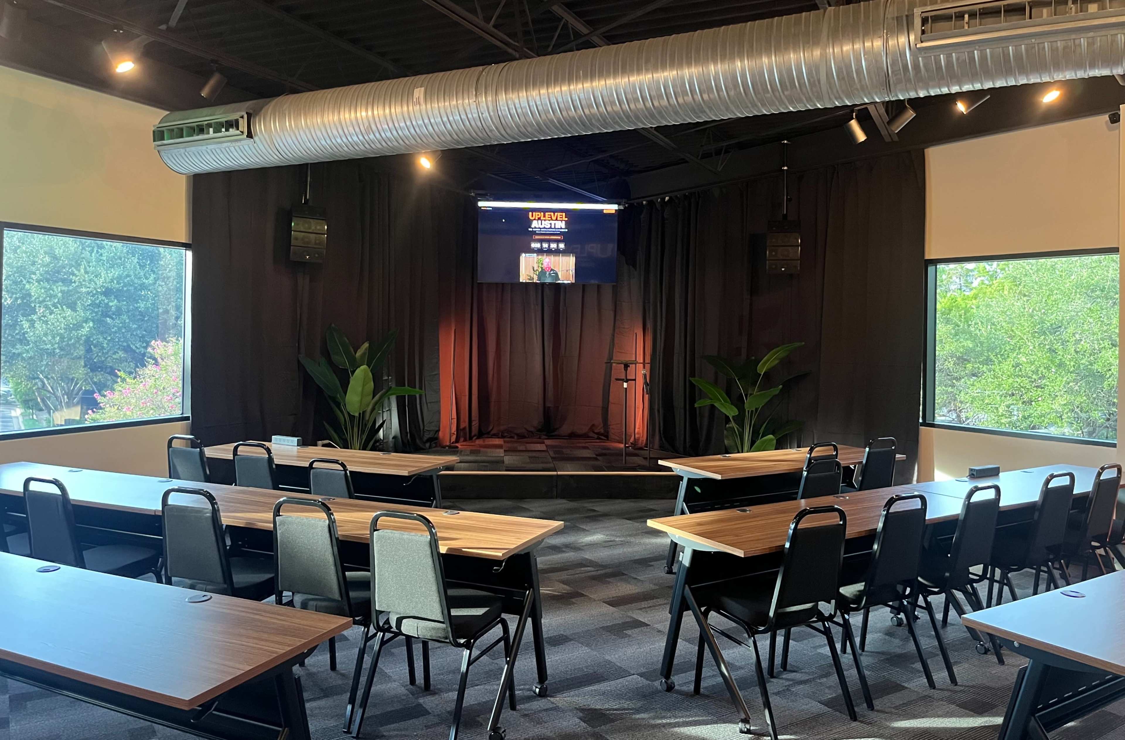 The image shows an indoor meeting room with rows of tables and chairs facing a stage area, complete with a microphone and a screen displaying text.