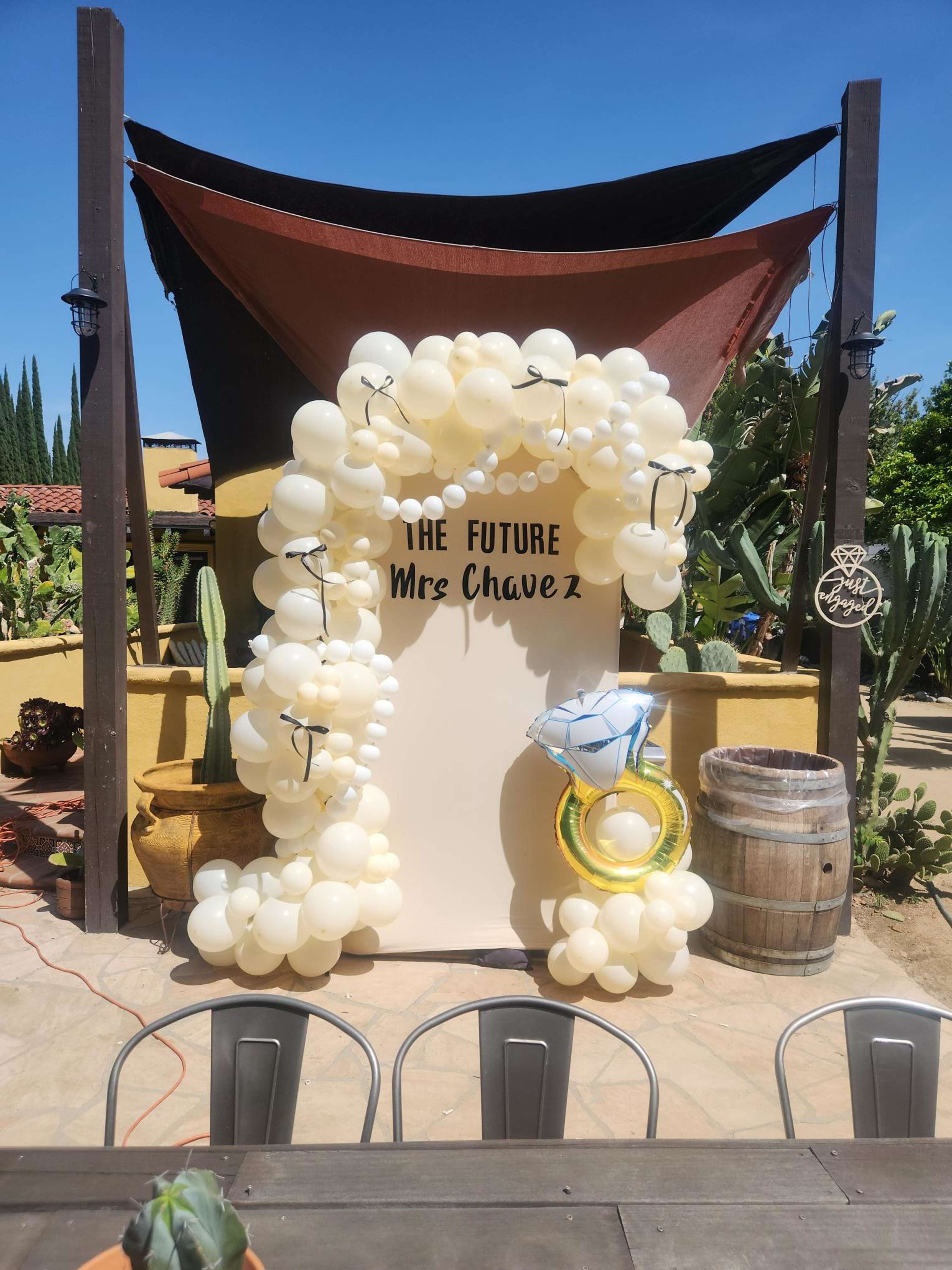 The scene features a decorative balloon arch with the text "Mrs. Chavez" set up in a rustic outdoor setting surrounded by cacti and barrels.