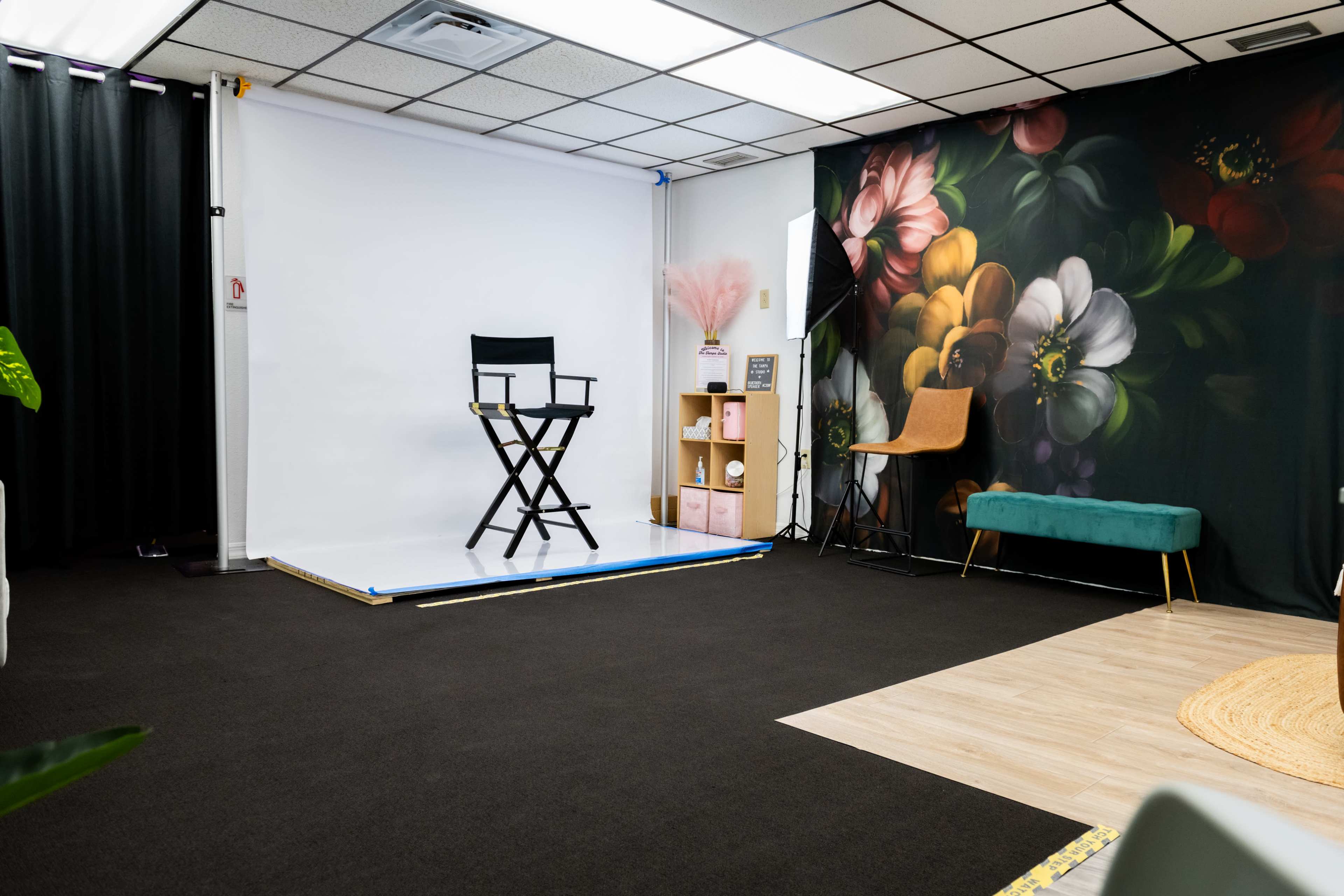 The image shows a photography studio with a white backdrop, a director's chair, and a floral mural on one wall alongside decorative furniture.