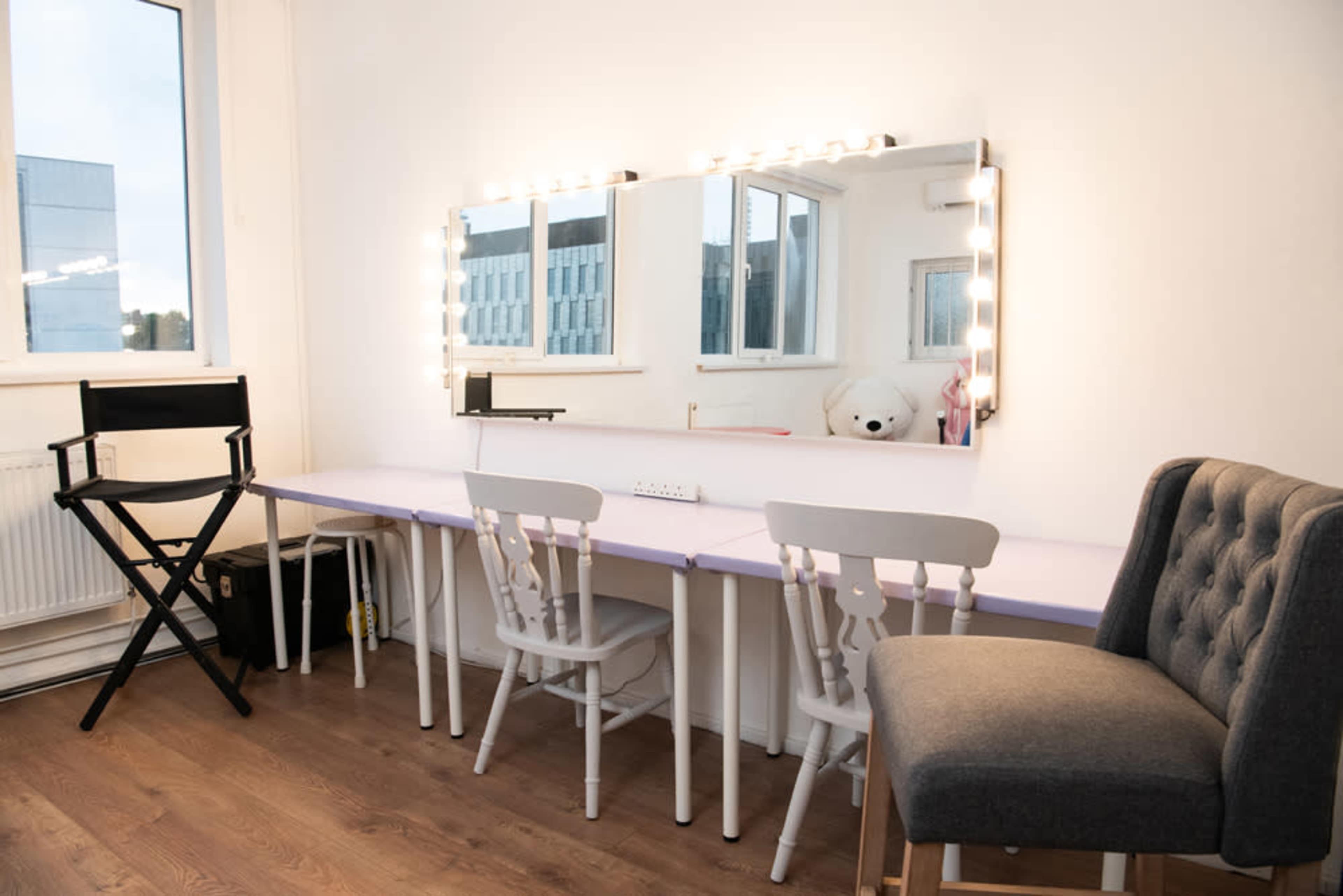 A well-lit vanity area features a large mirror with light bulbs, two white chairs, a gray upholstered chair, and a black director's chair, all set against a white wall.