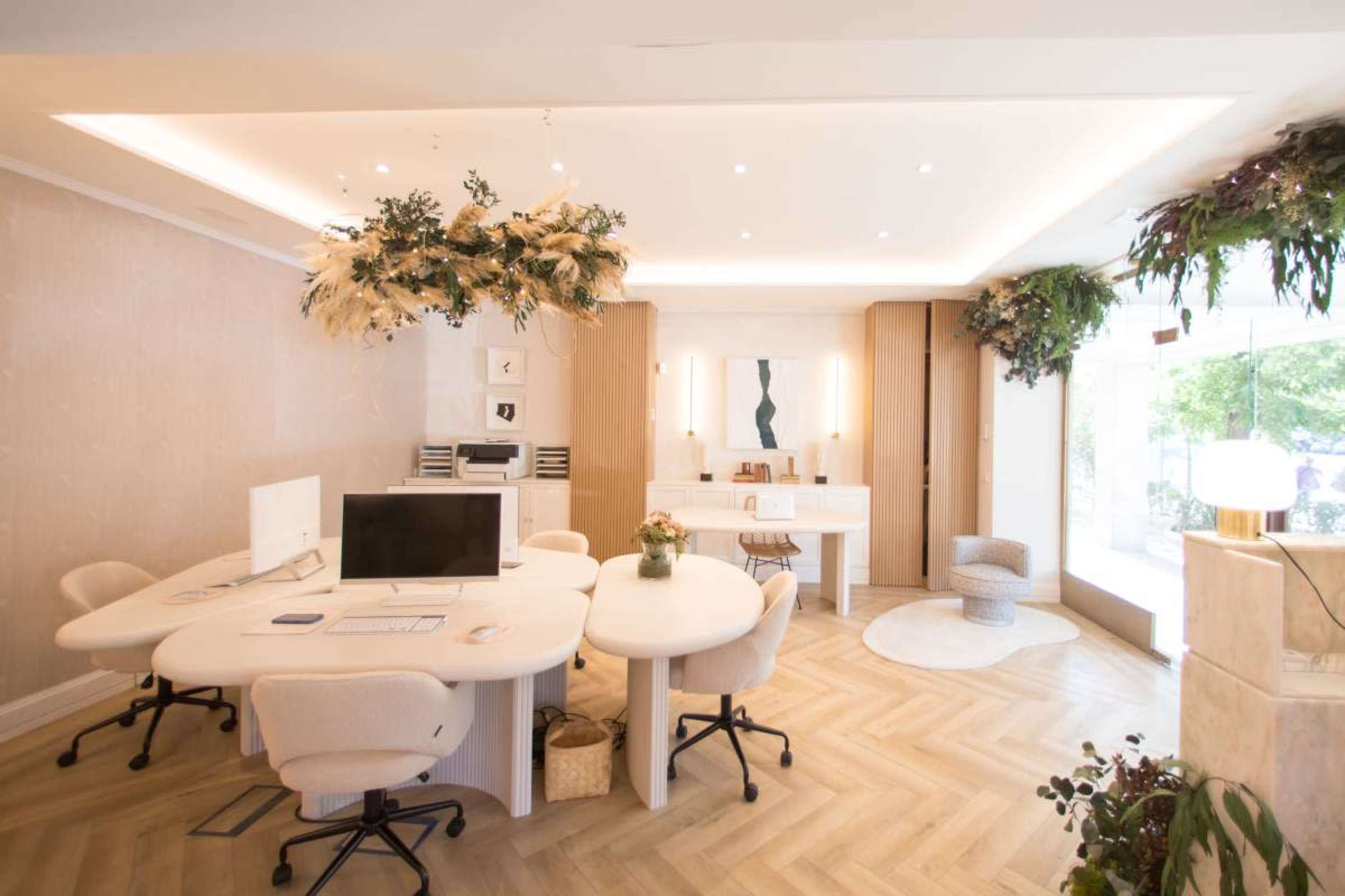 The image shows a modern office space with circular desks, light wood flooring, and decorative plants hanging from the ceiling.