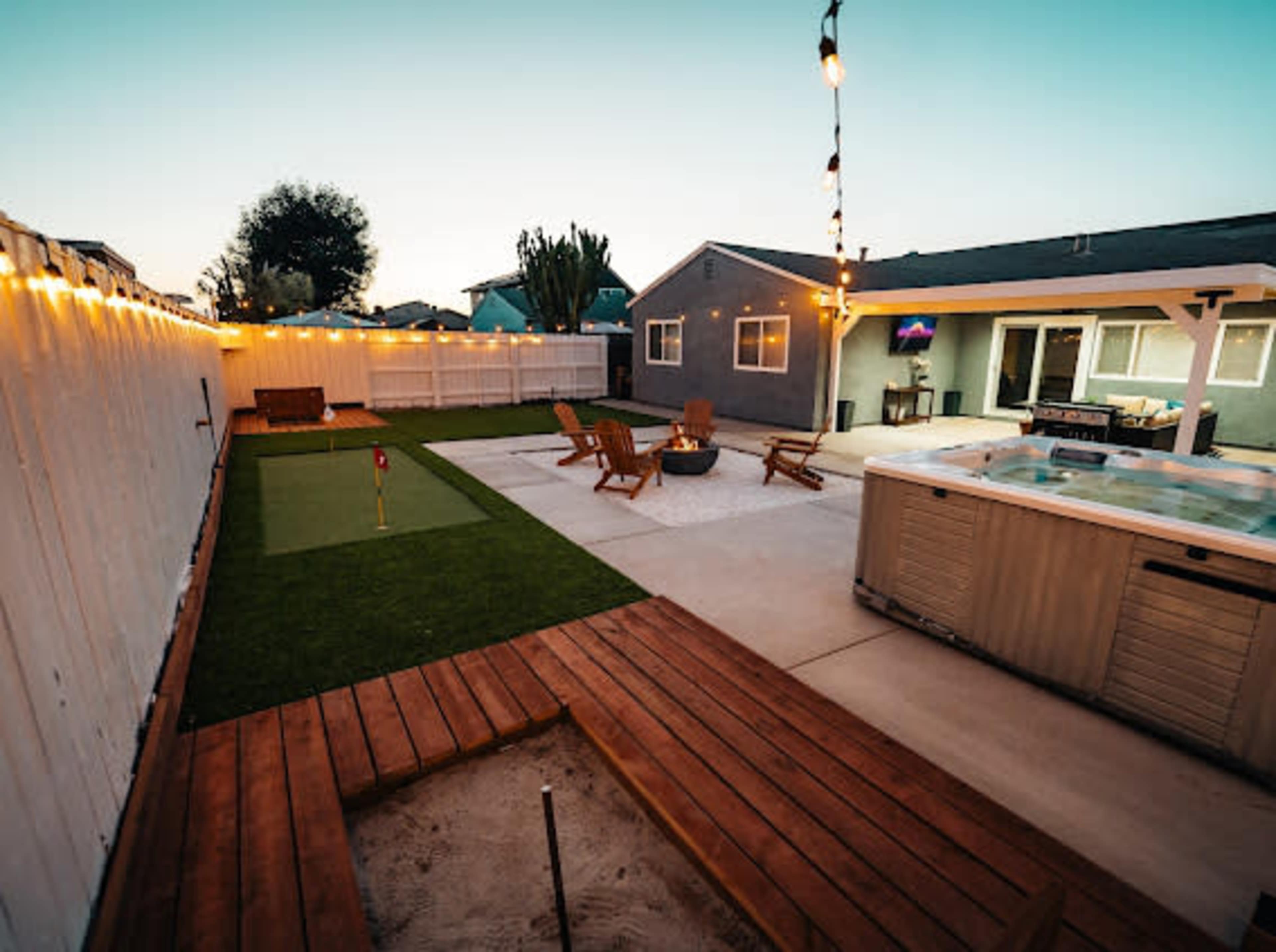 The backyard features a putting green, a fire pit surrounded by seating, a hot tub, and decorative string lights along a white fence.