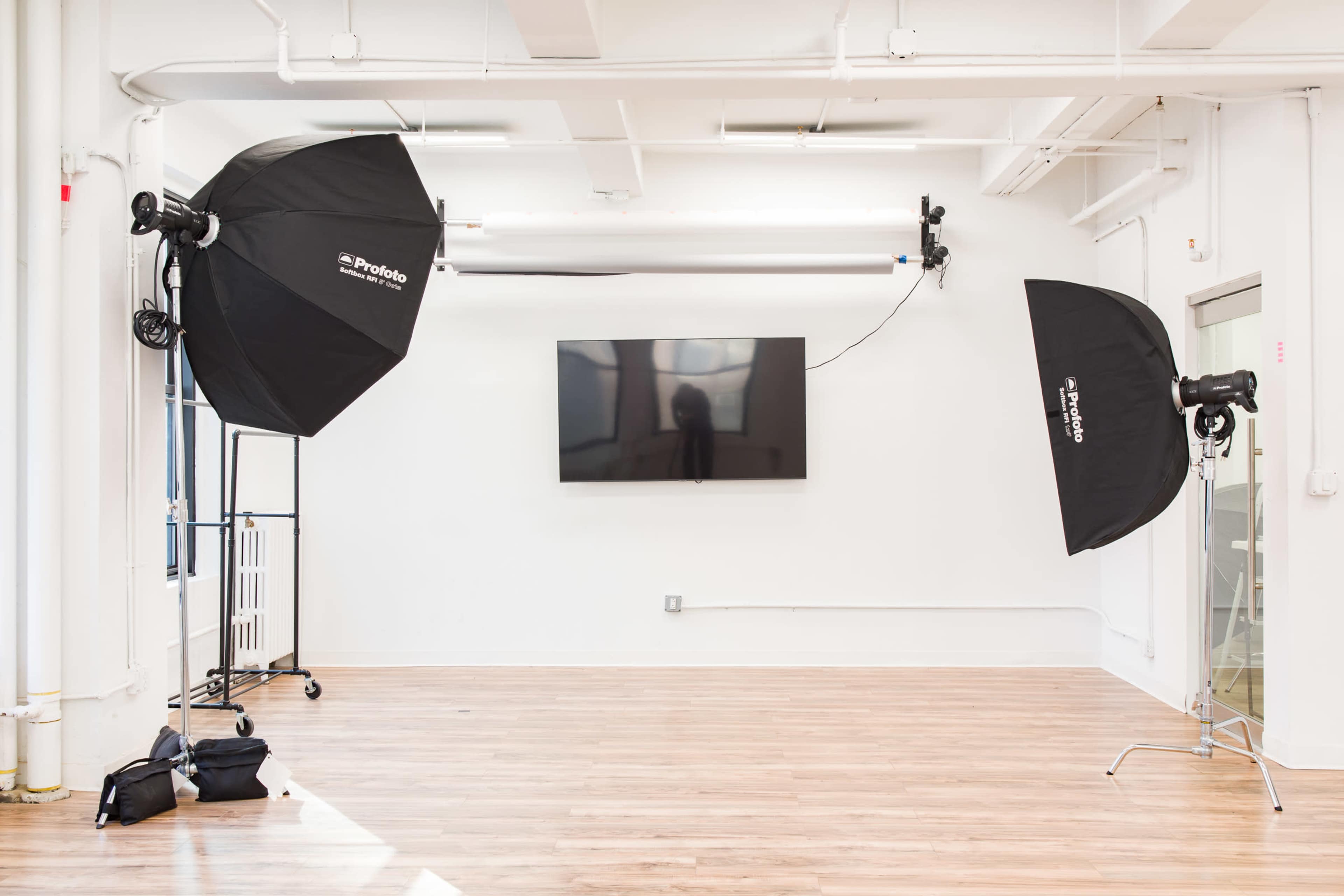 The image shows a bright, empty studio space with two photography light umbrellas on stands and a large flat-screen TV mounted on the wall.
