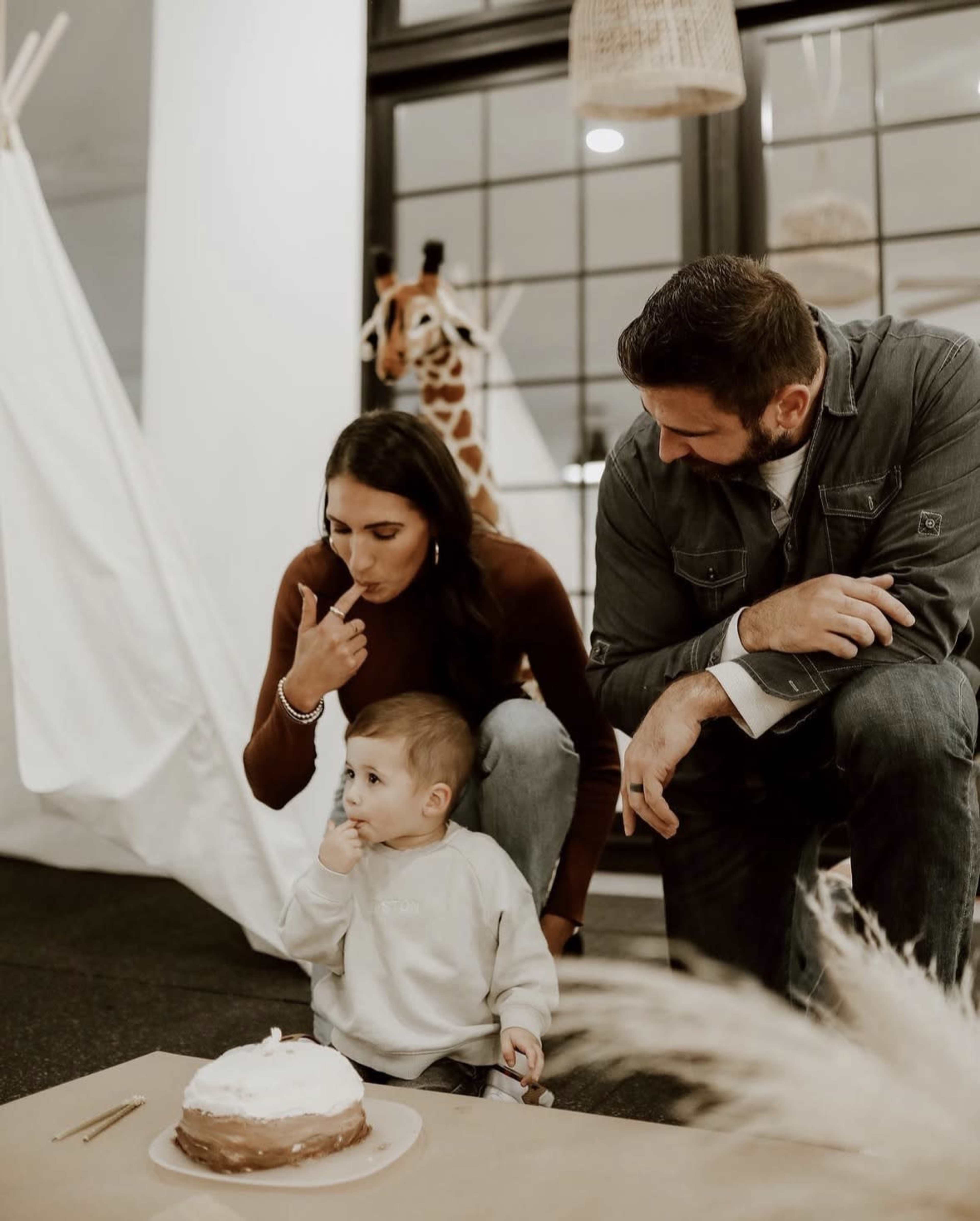 A woman and a man kneel beside a toddler, who is sitting in front of a cake on a table in a decorated indoor space.