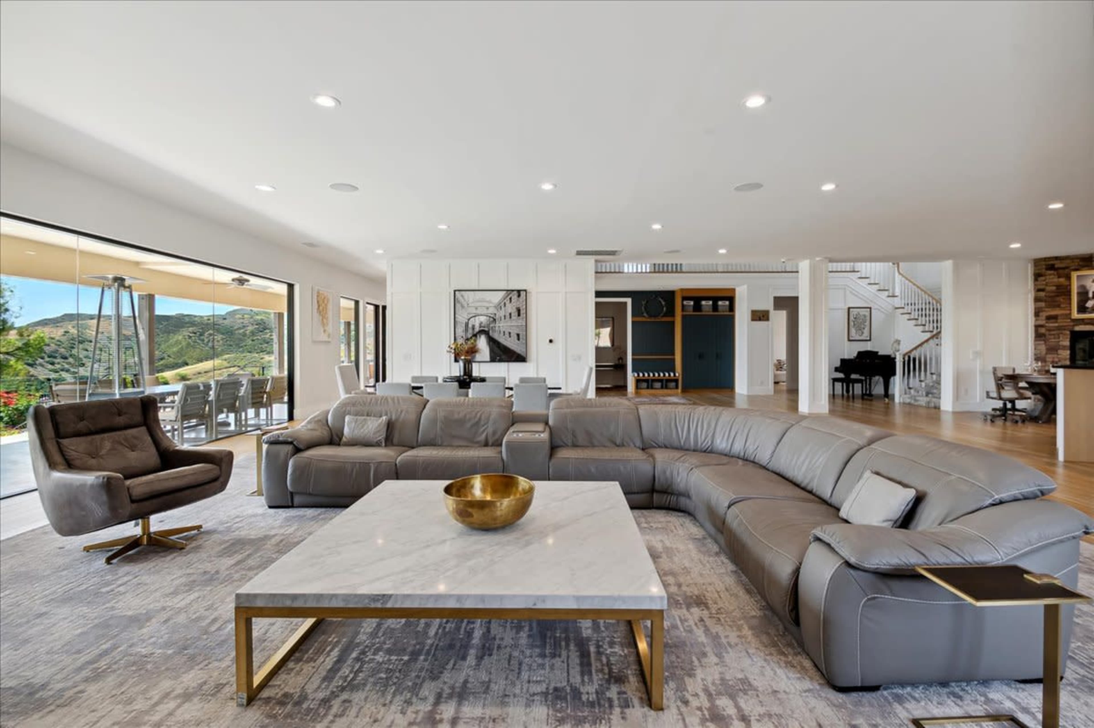A spacious modern living room features a curved gray leather sectional sofa, a marble coffee table, and large glass doors that open to an outdoor patio with a view.