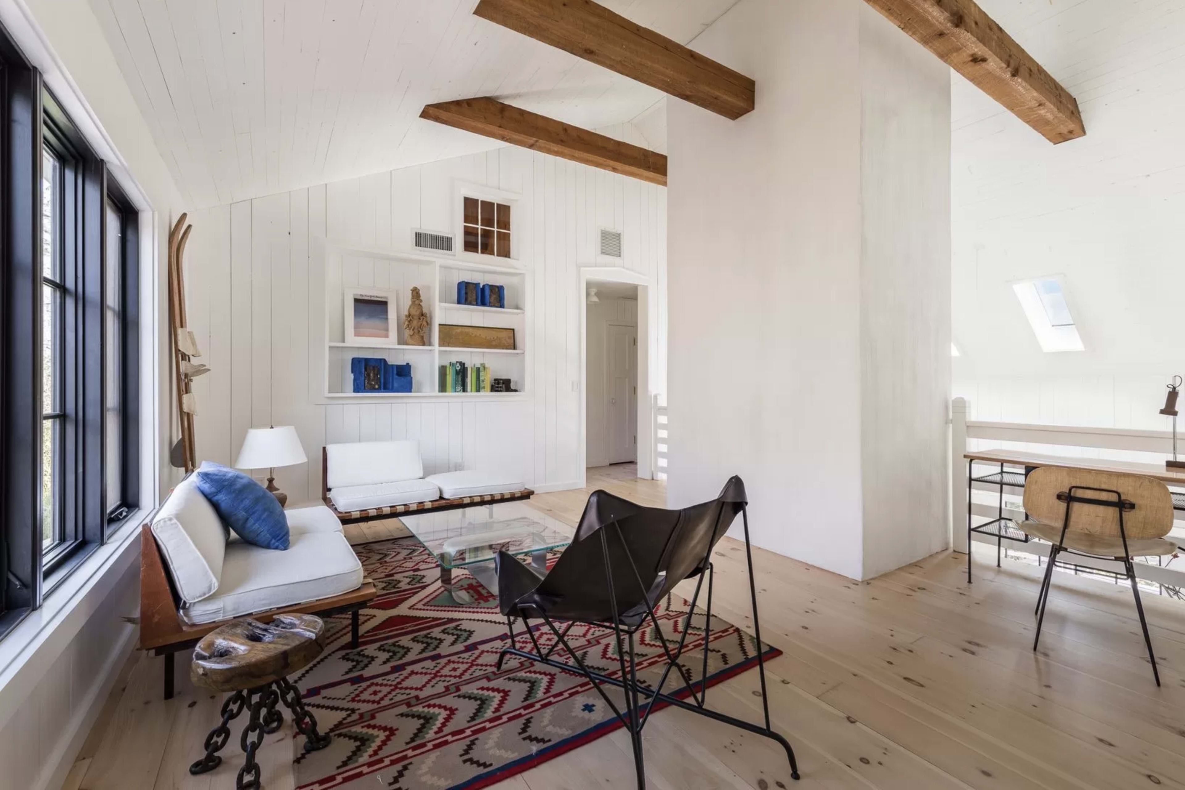 A modern living space with white walls, wooden beams, a glass coffee table, a woven rug, and a mix of seating options, including a sofa and a folding chair.